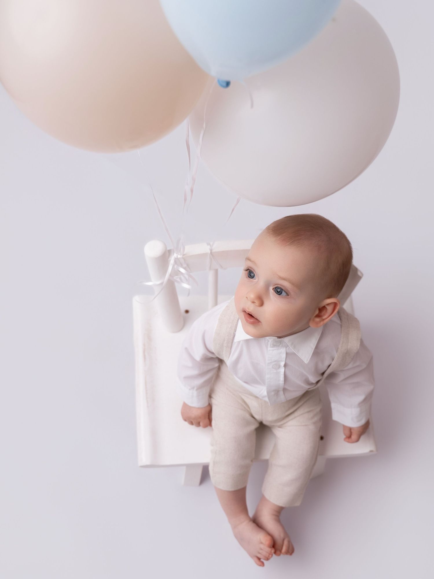 Baby boy in white shirt and beige pants sits on white box, gazing up at pastel balloons against white background.
