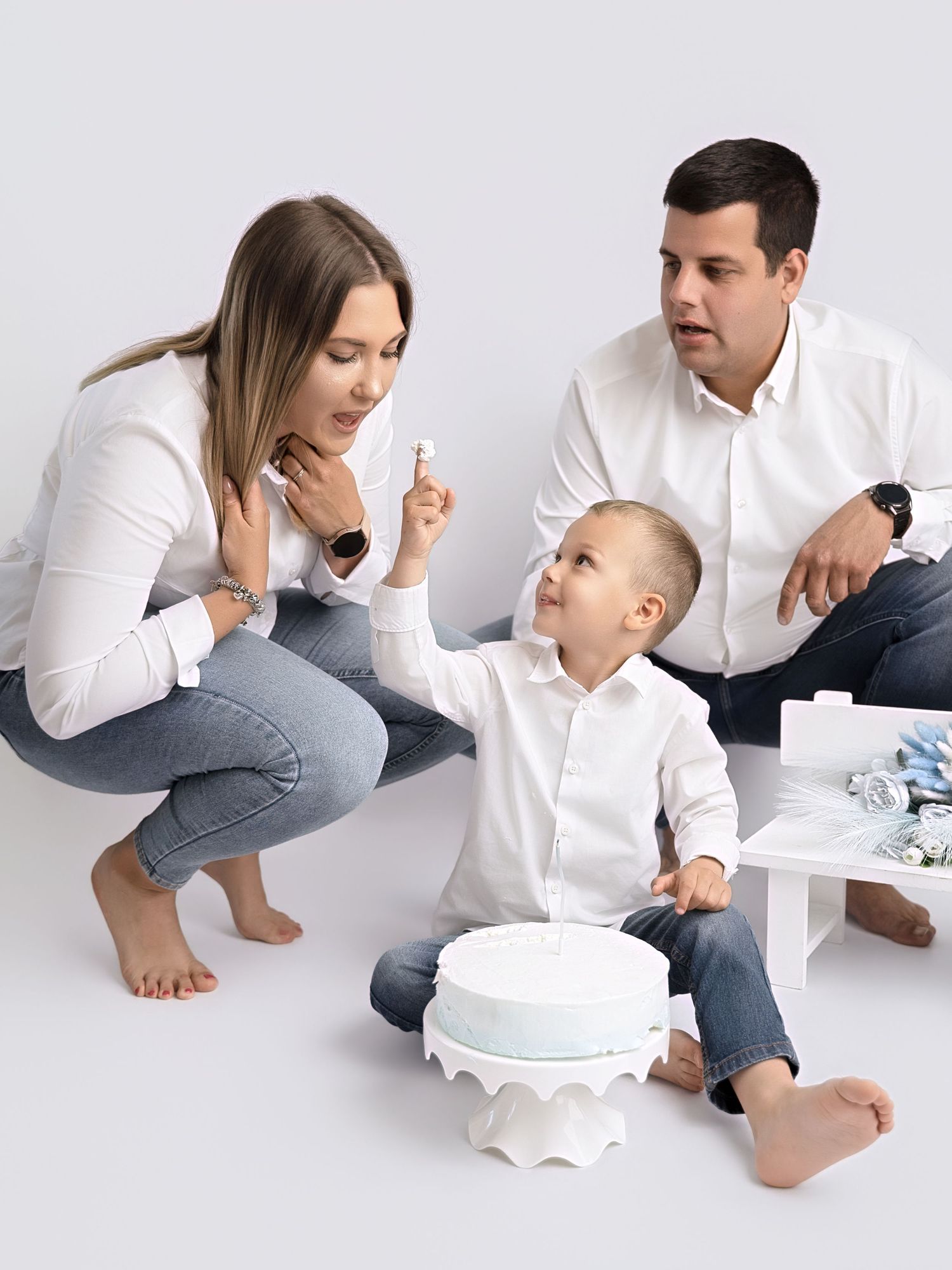 Family celebrating toddler's first birthday with white cake in a bright studio photo shoot.