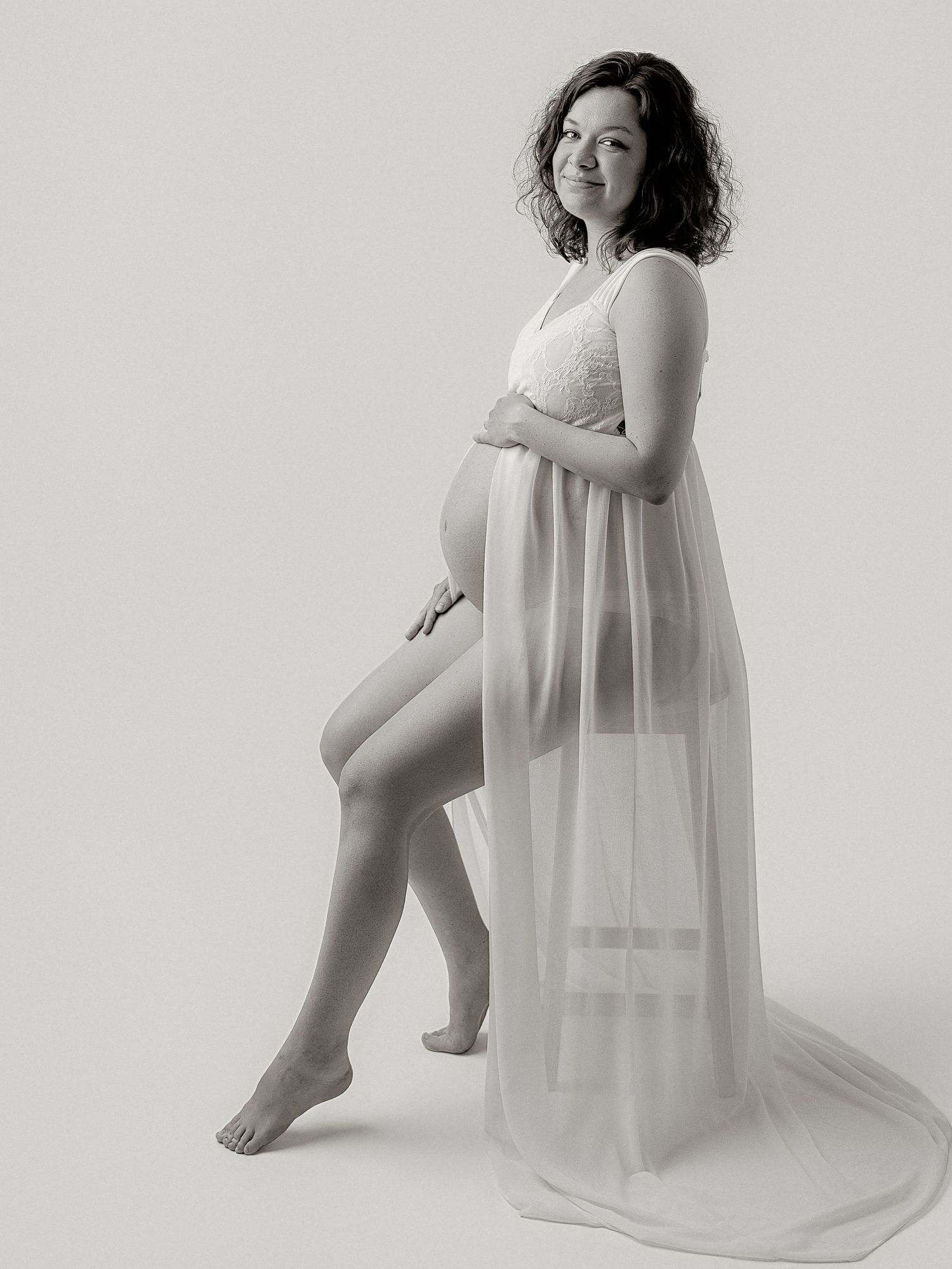Pregnant woman in white sheer maternity gown posing gracefully in a black and white studio portrait photo.