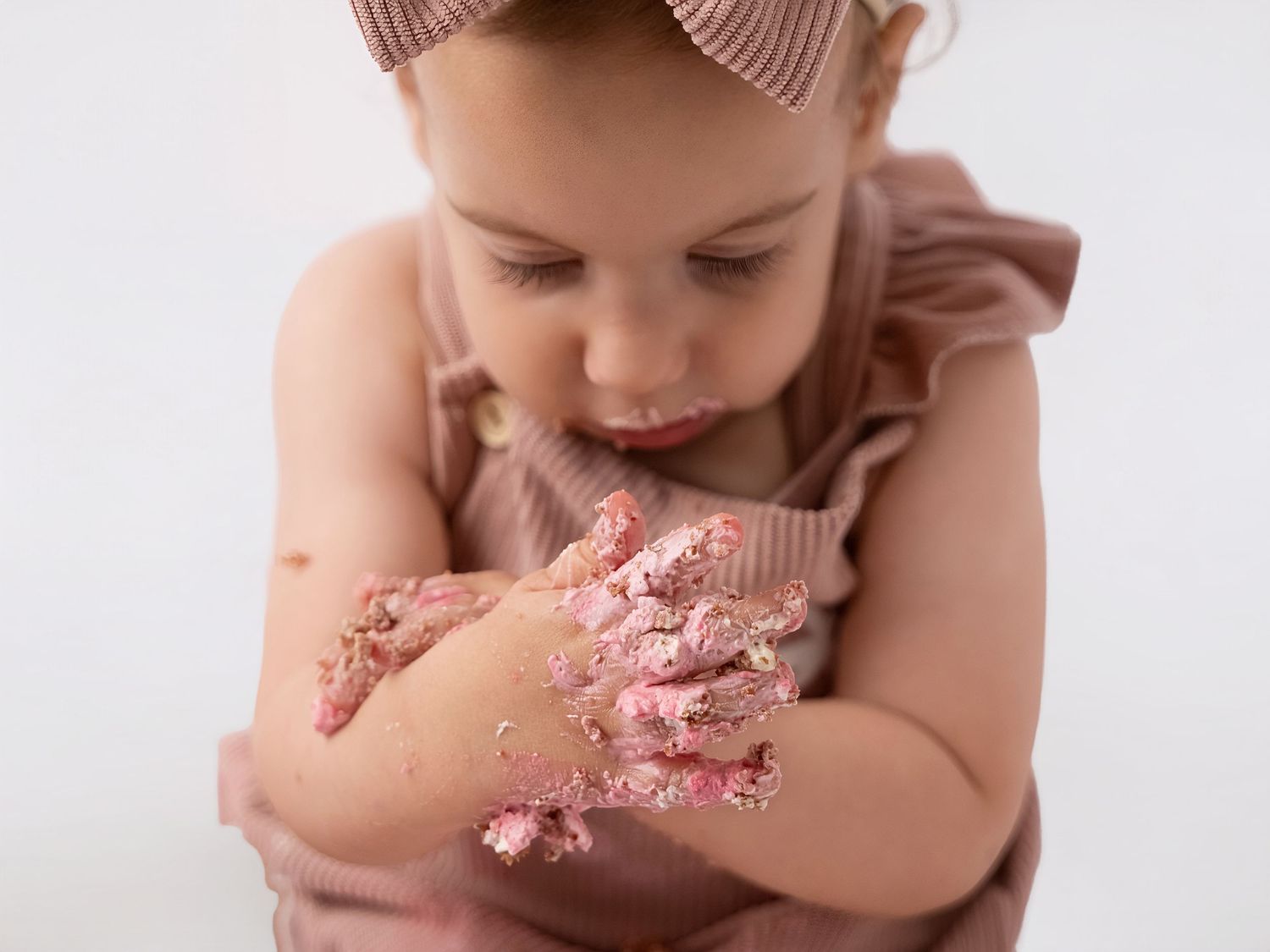 Toddler girl in pink romper with cake smash on her hands during first birthday photoshoot.