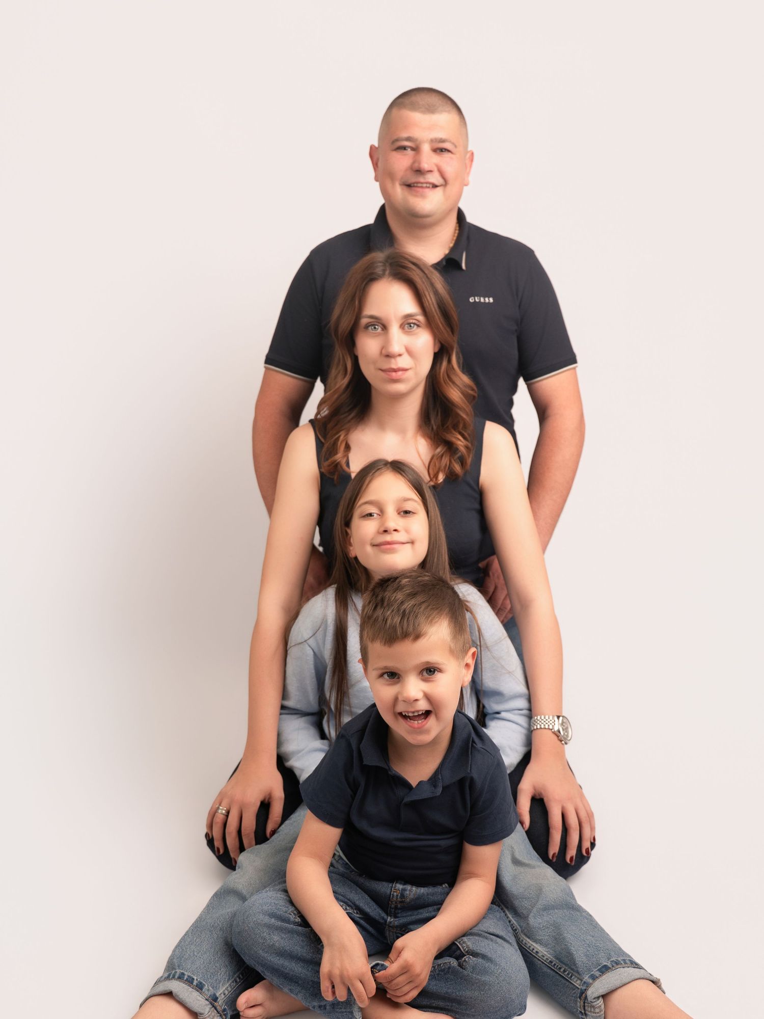 A happy family of four posing for a studio portrait, with parents and two young children arranged in a vertical stack.