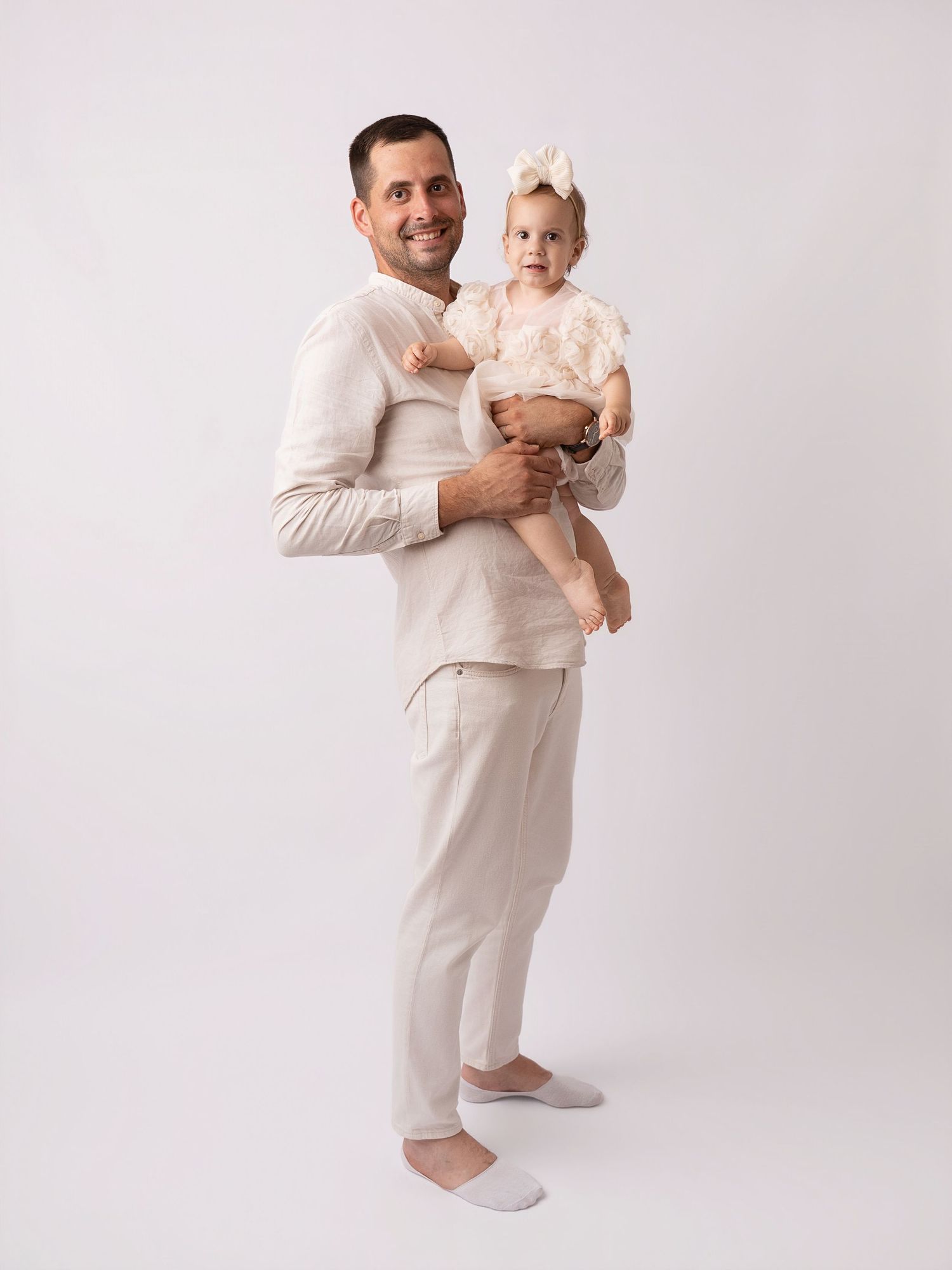 A smiling man in beige linen outfit holds a baby girl with a white bow on a white background.