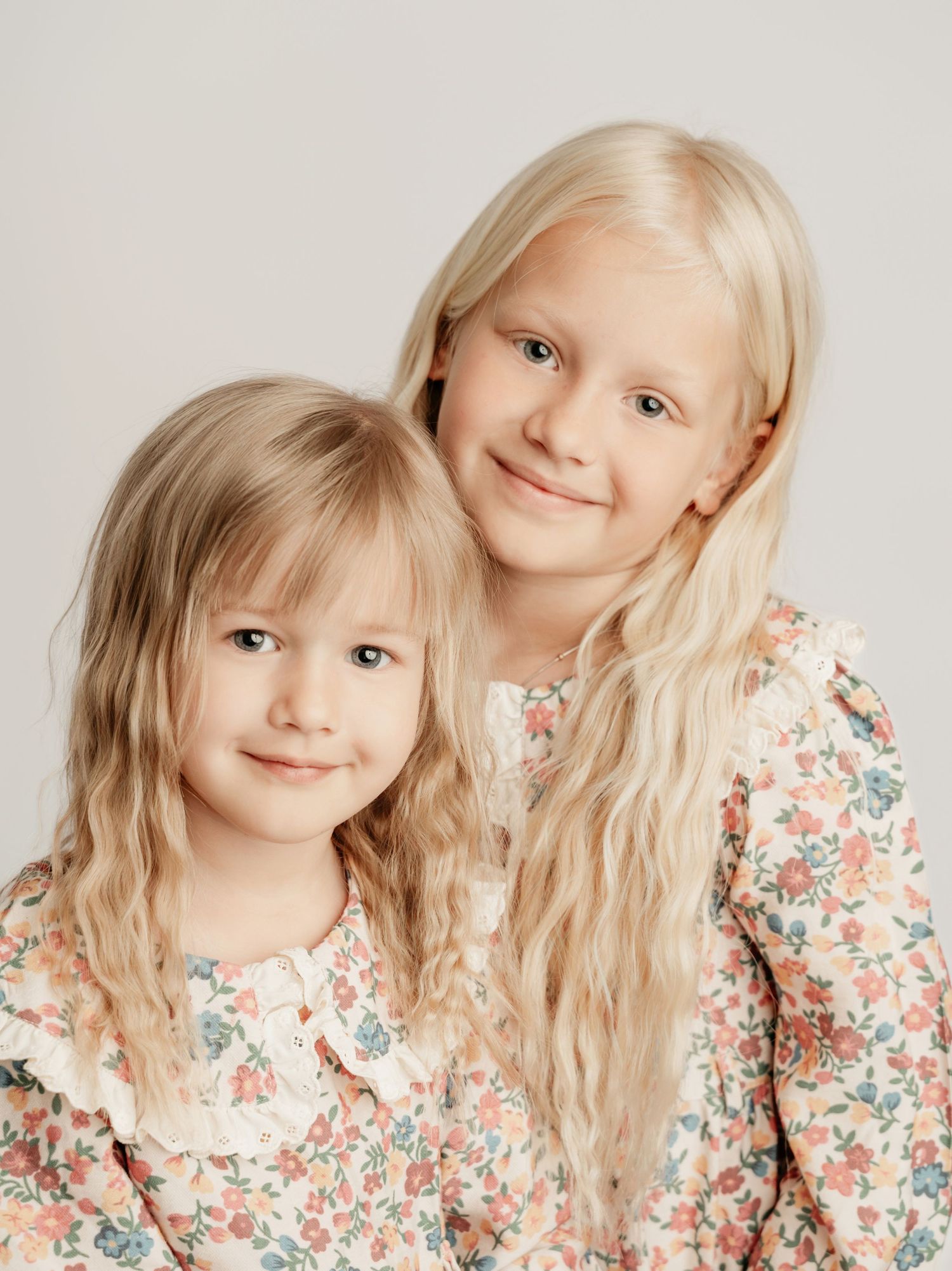 Two blonde sisters in matching floral dresses posing together in soft light studio children portrait.
