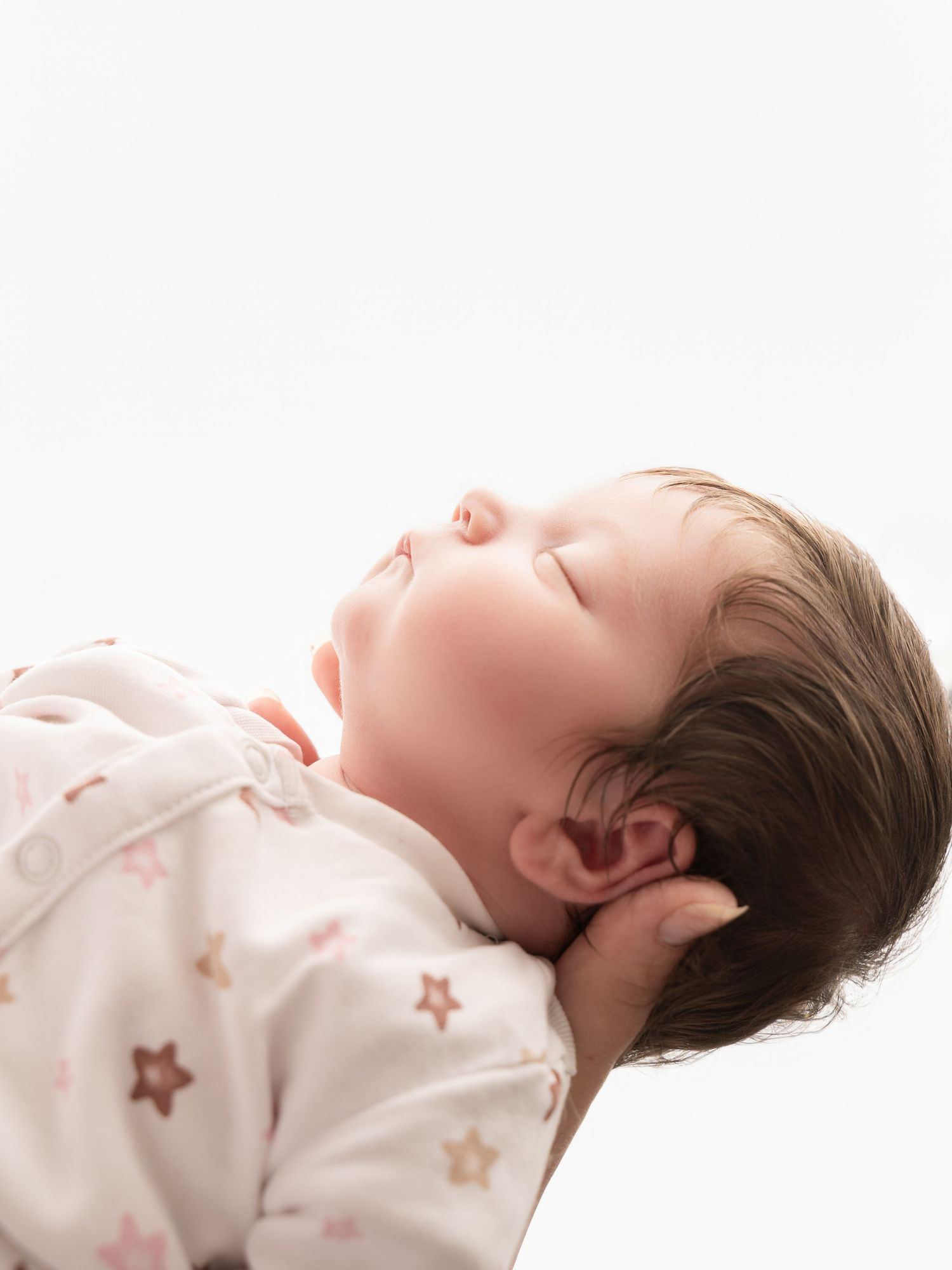 Side profile of sleeping newborn baby with dark hair in star-print onesie against white background.