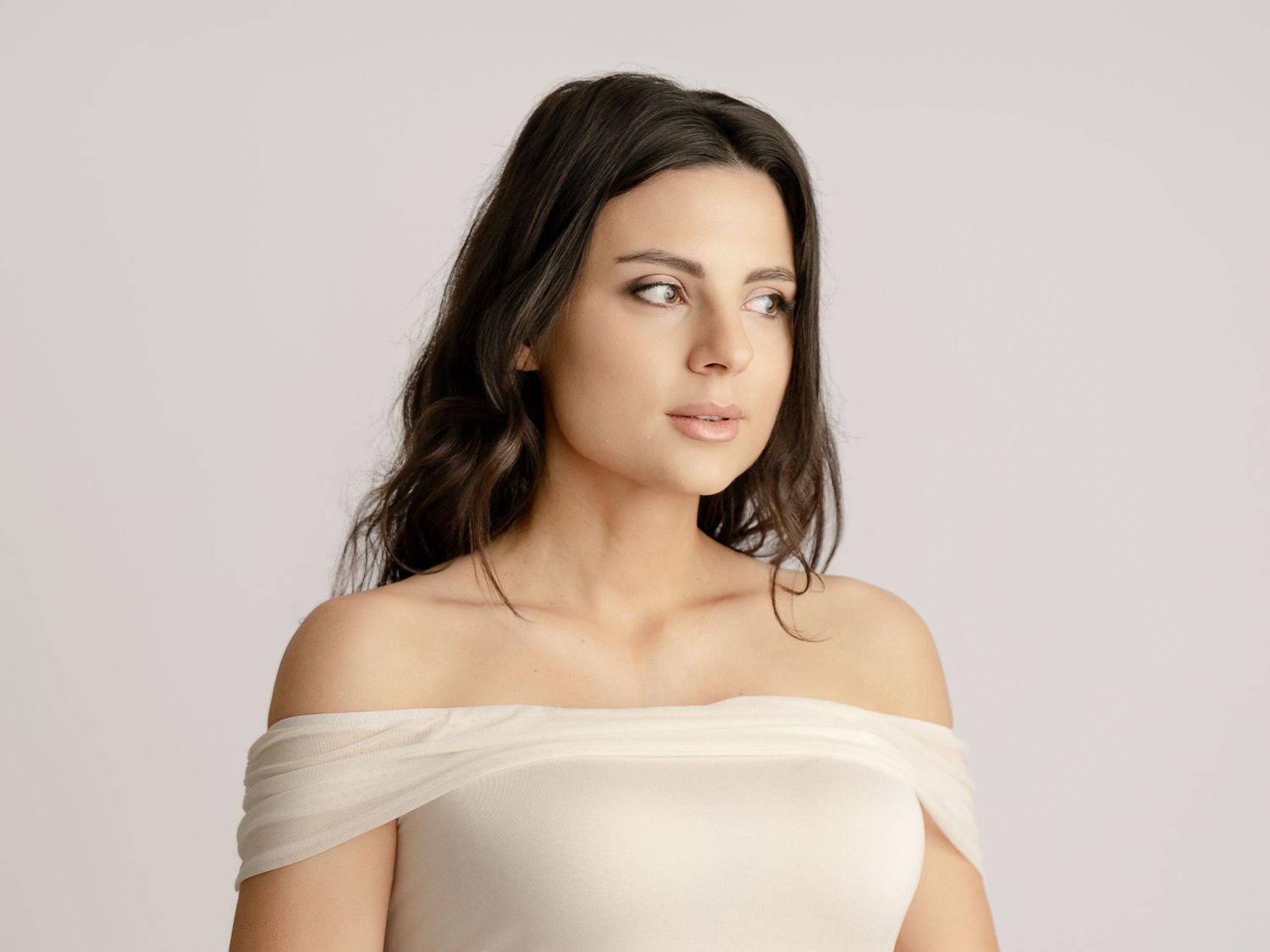 Brunette woman in white off-shoulder top poses elegantly against neutral gray background.