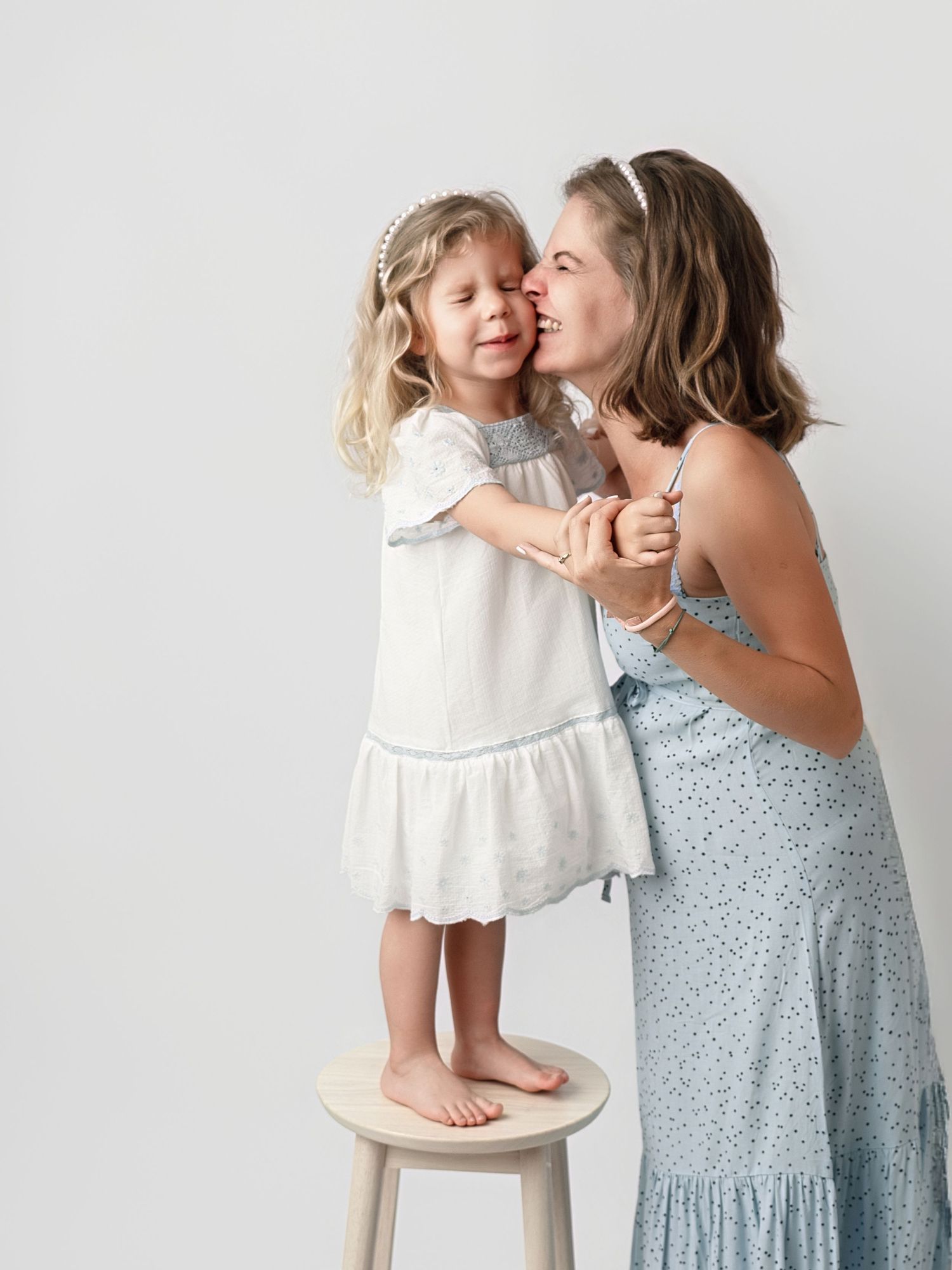 A woman in a blue floral dress kisses a young girl in a white dress standing on a stool against a light background.