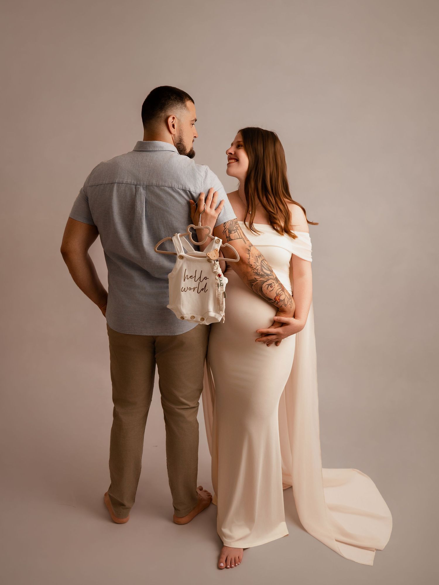 Couple posing for maternity photo in studio, woman in white gown holding tiny baby outfit labeled hello world.