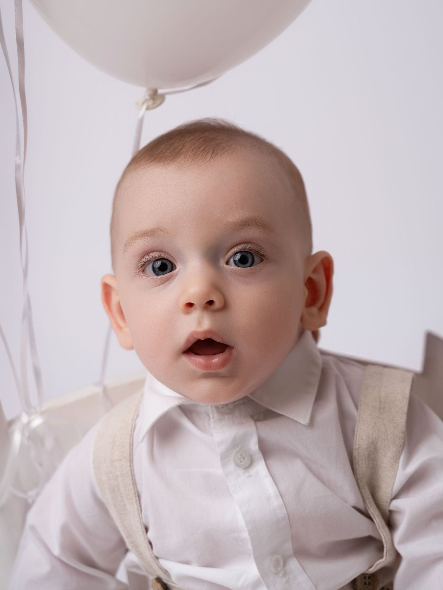 Adorable baby boy in white shirt and suspenders with wide eyes and open mouth next to a white balloon.