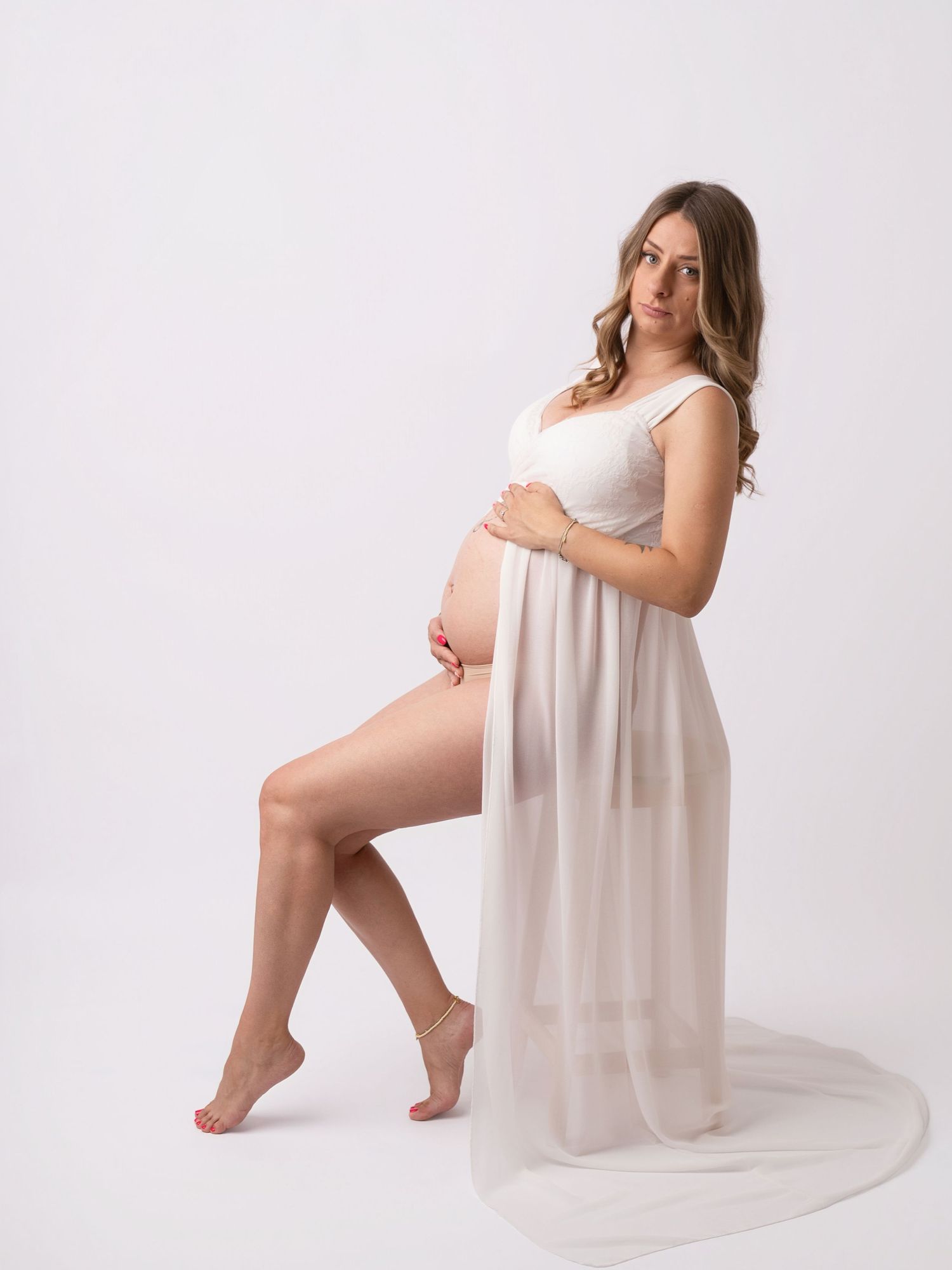 Pregnant woman in white sheer maternity gown posing gracefully against a white studio background.