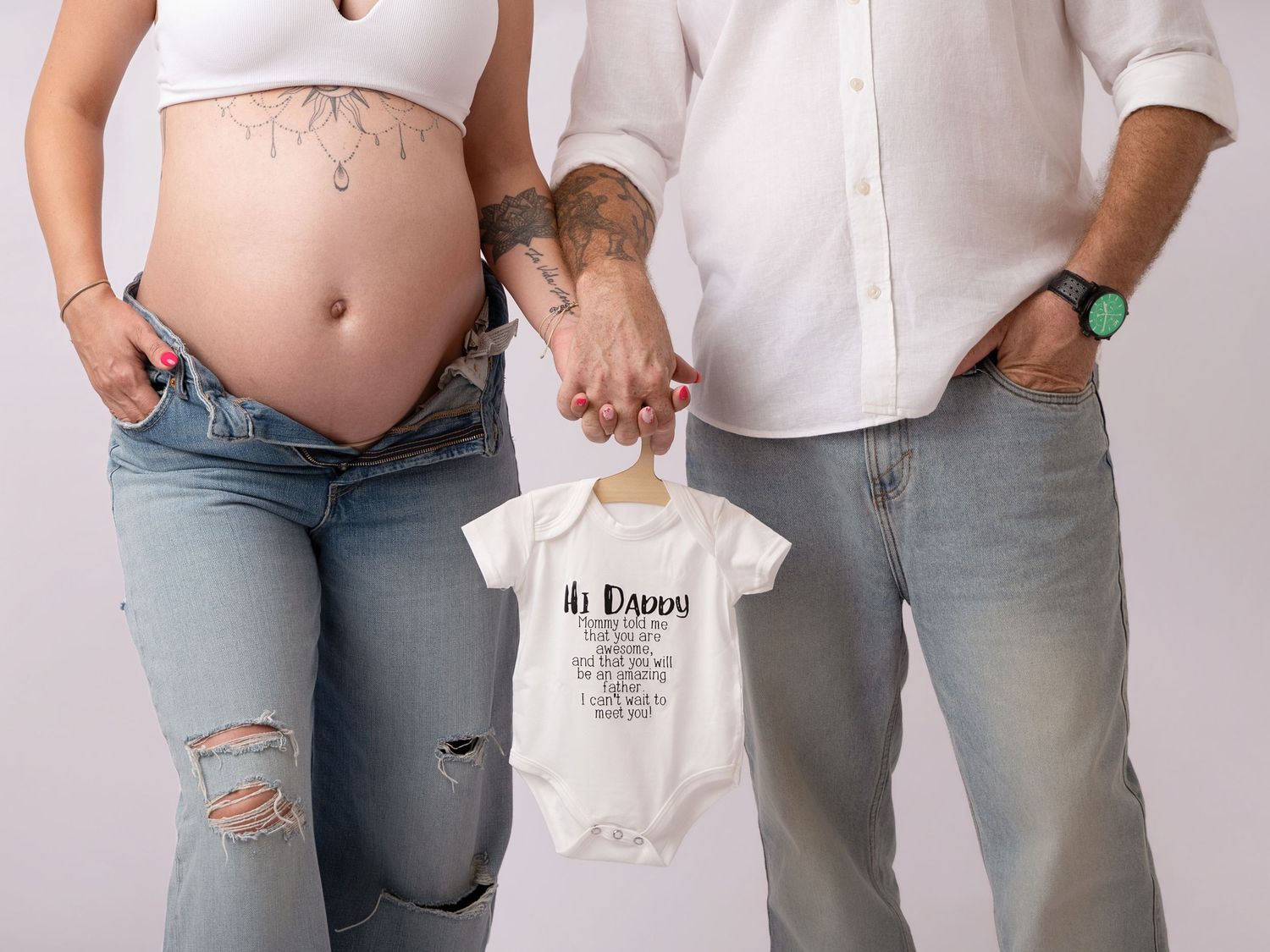 Pregnant couple holding a white Hi Daddy baby onesie, maternity announcement photo with tattoos and casual attire.