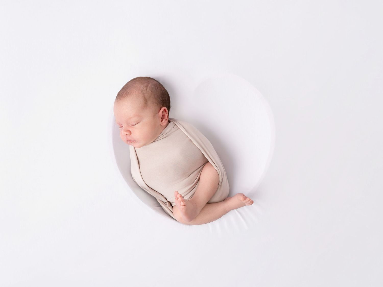 Peaceful newborn baby sleeping curled up in a white bowl prop, wrapped in a soft beige onesie on white background.