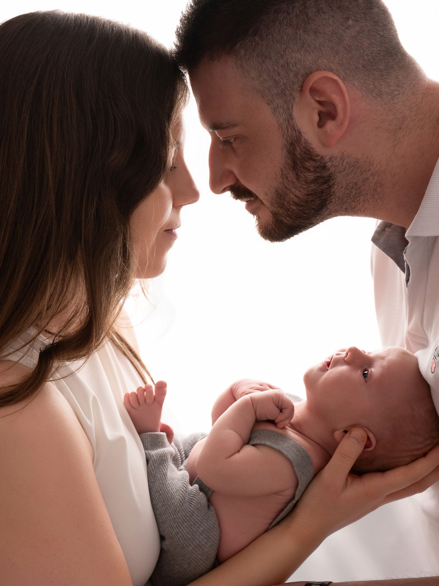 Two parents lean in close, tenderly holding their newborn baby in a bright, intimate family portrait.