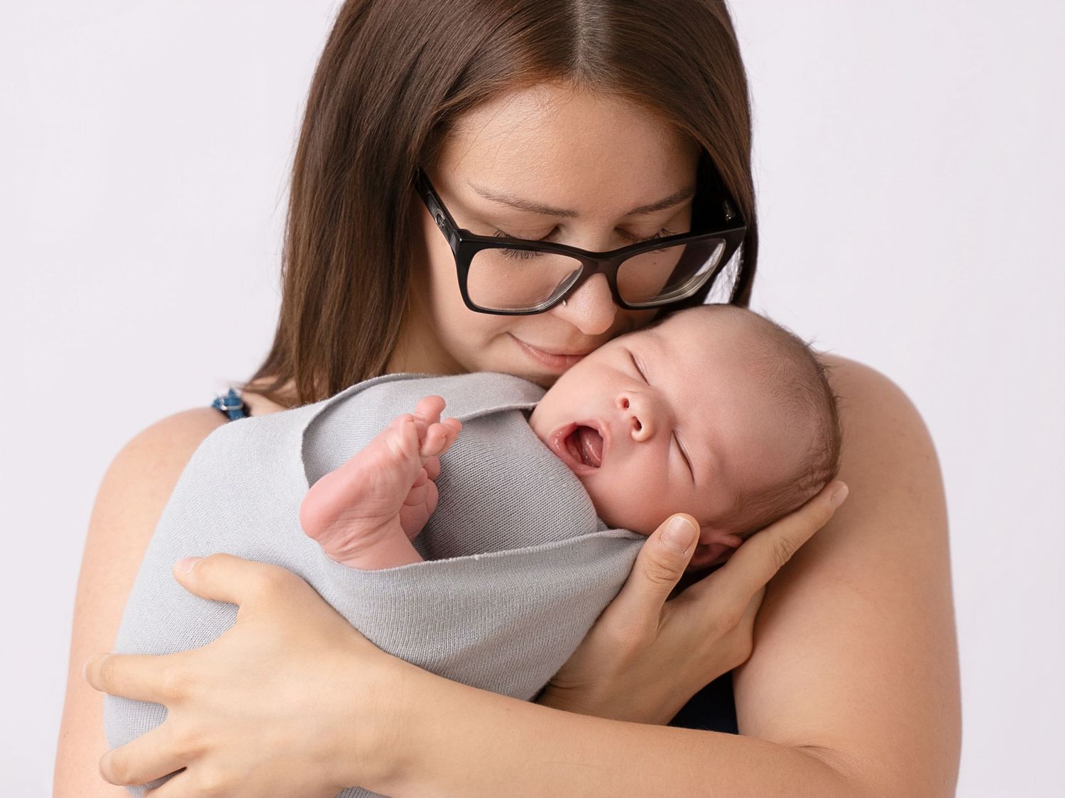 A mother with glasses tenderly holds her swaddled newborn baby against her chest on a white background.