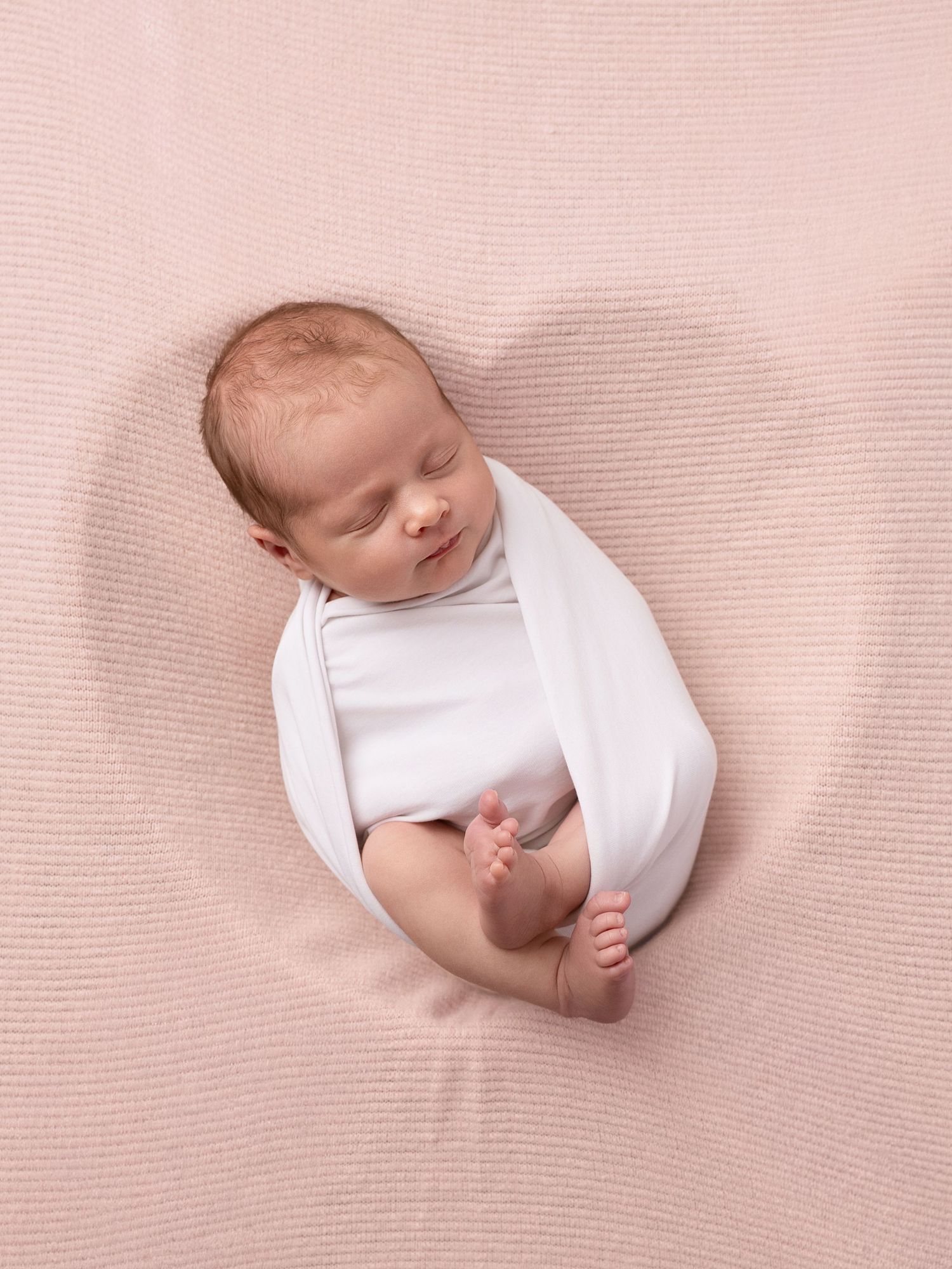 Newborn baby swaddled in white wrap sleeping peacefully on a soft pink textured blanket.