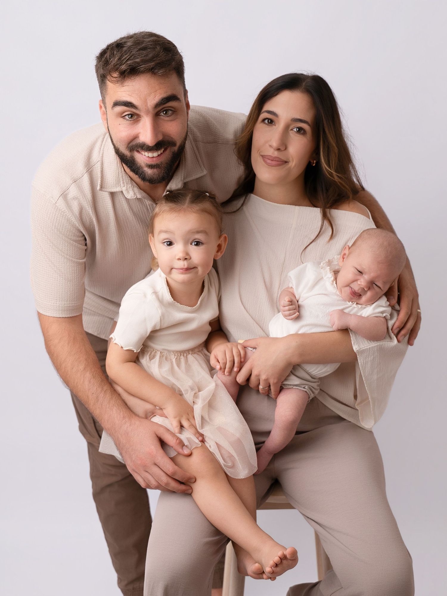 A smiling family of four poses in a studio, with parents holding a toddler girl and a newborn baby, dressed in neutral tones.
