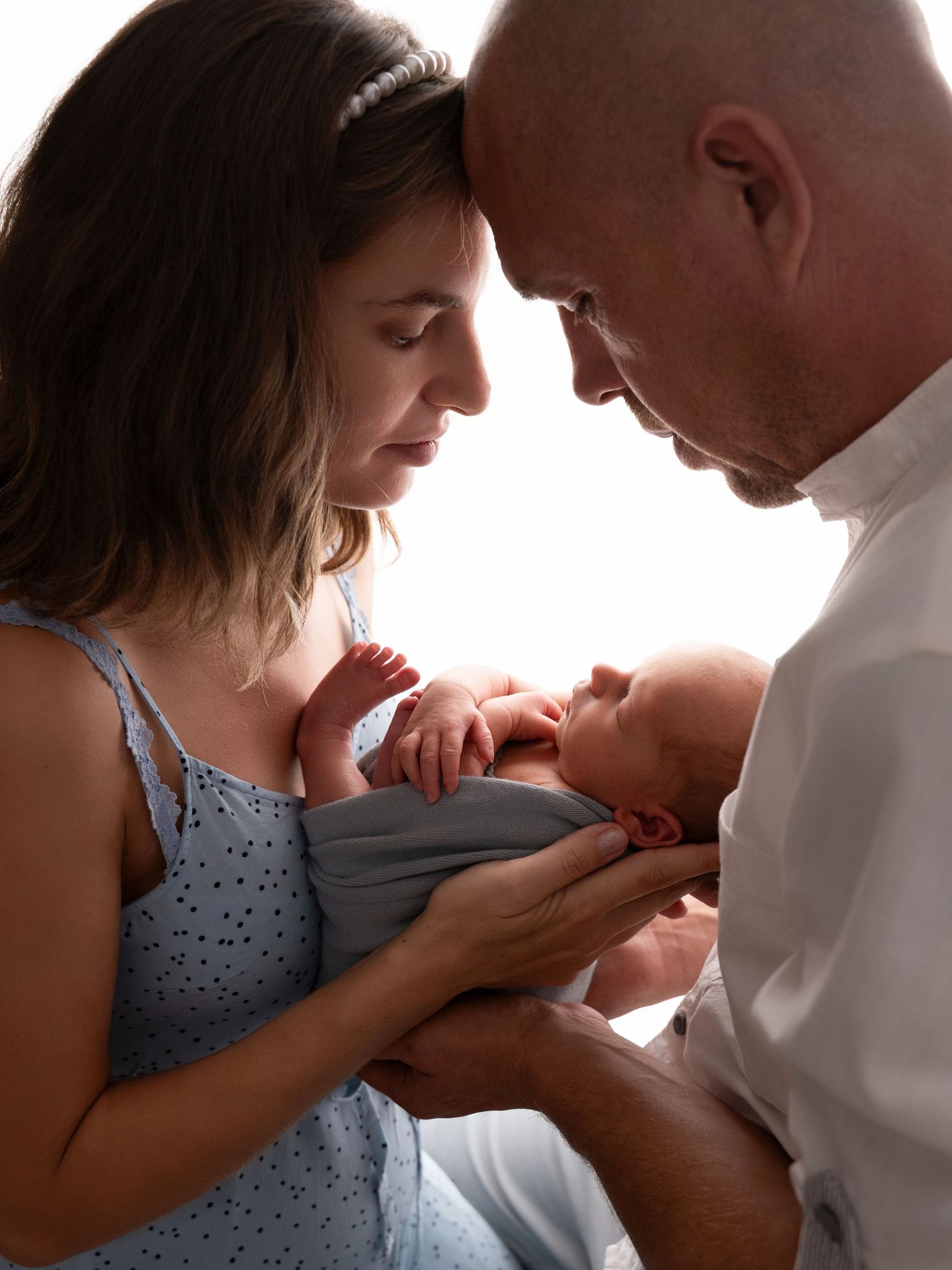 New parents tenderly hold their sleeping newborn baby together, foreheads touching in an intimate family portrait.