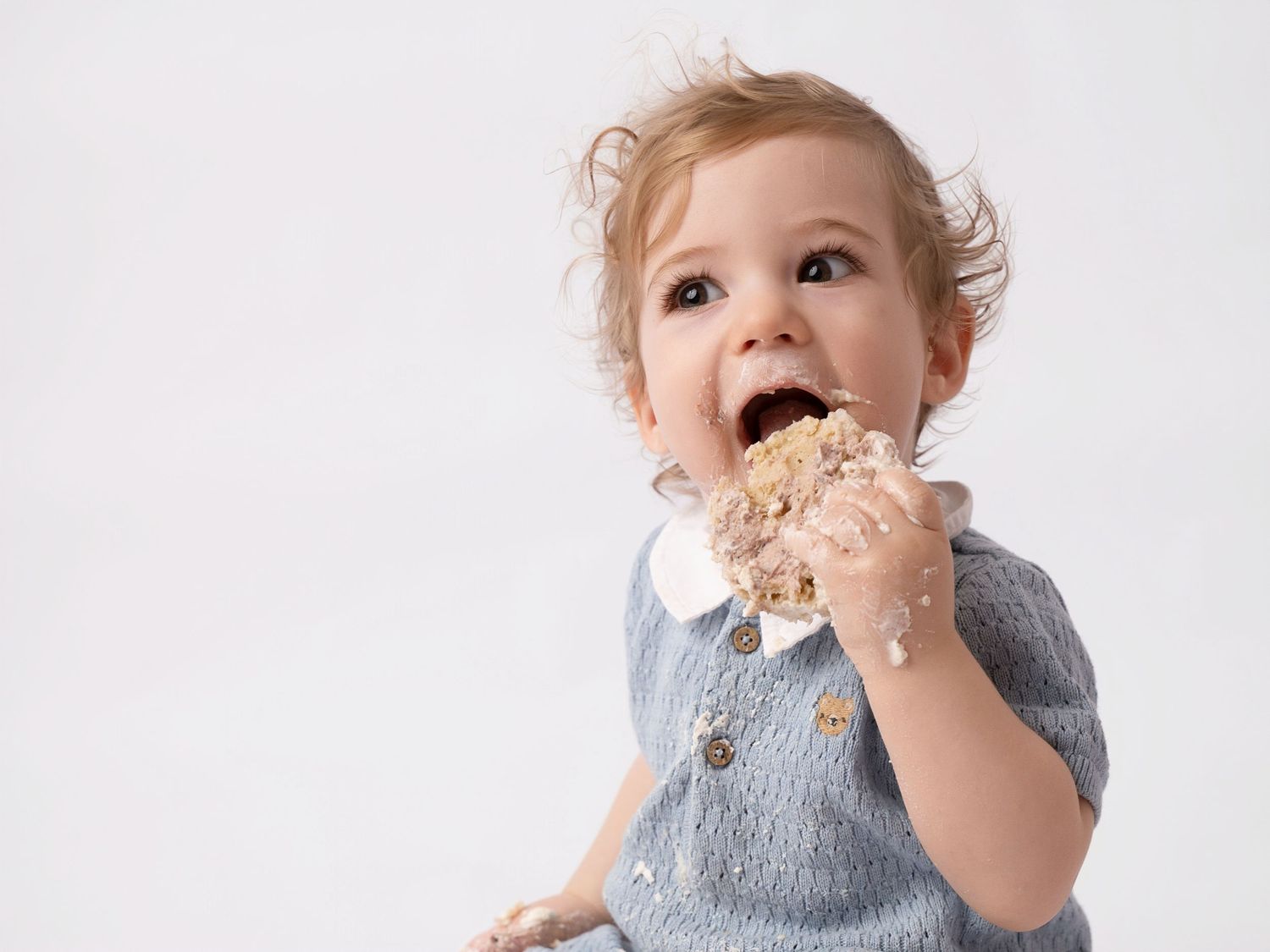 A cute toddler in a blue knit outfit eagerly eating a piece of cake with frosting on their hands and face.