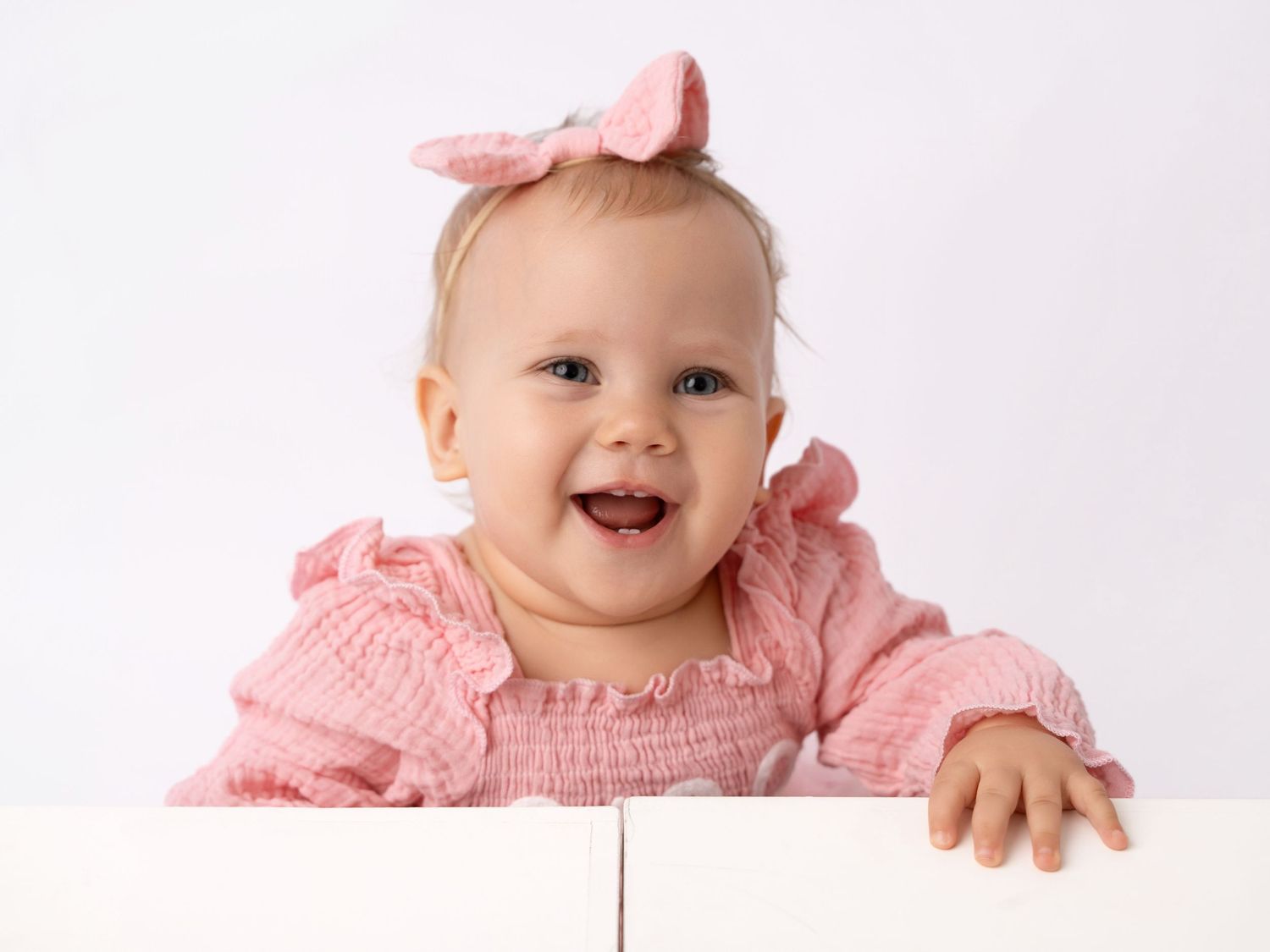 Smiling baby girl in pink ruffled outfit and bow headband peeking over white surface against light background.
