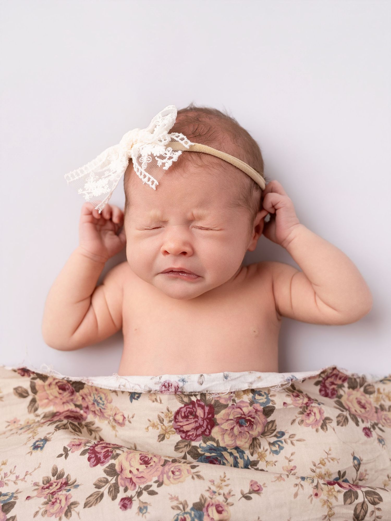 Newborn baby girl crying with hands raised, wearing a lace bow headband, wrapped in a floral blanket.