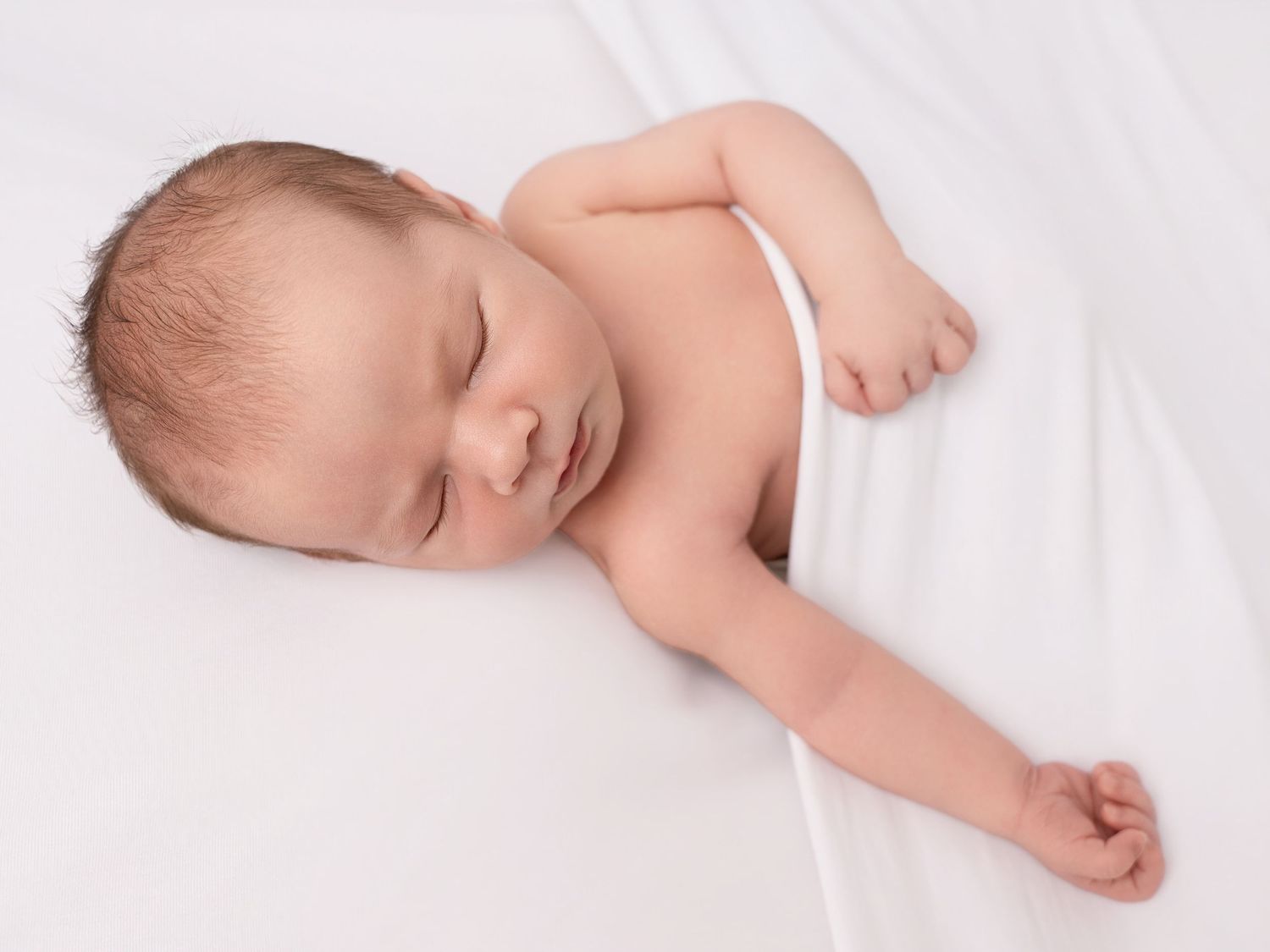 Peaceful newborn baby sleeping on white blanket, eyes closed, tiny hands relaxed in serene portrait.