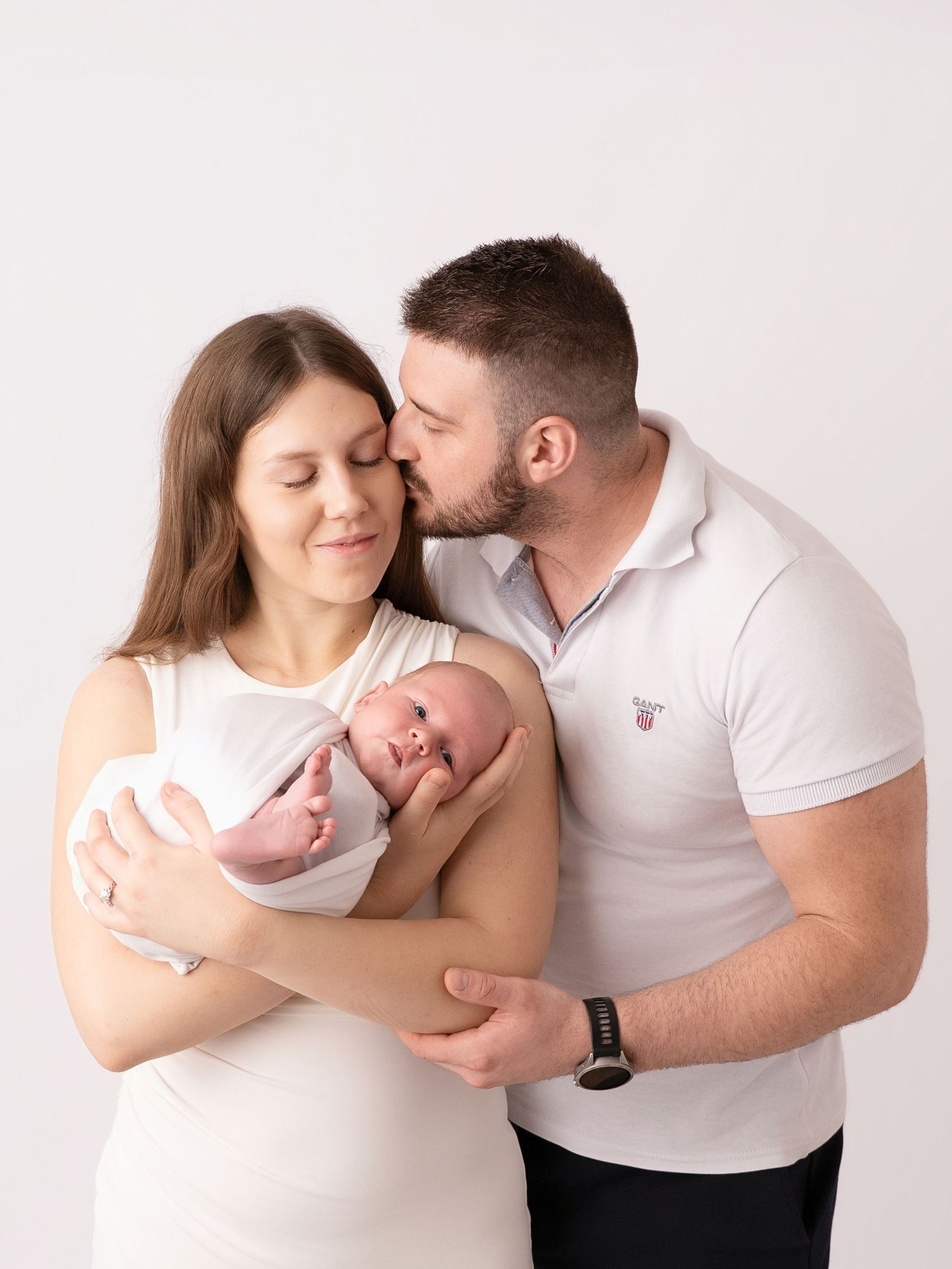A couple in white outfits tenderly holds their newborn baby, with the man kissing the woman on the forehead.