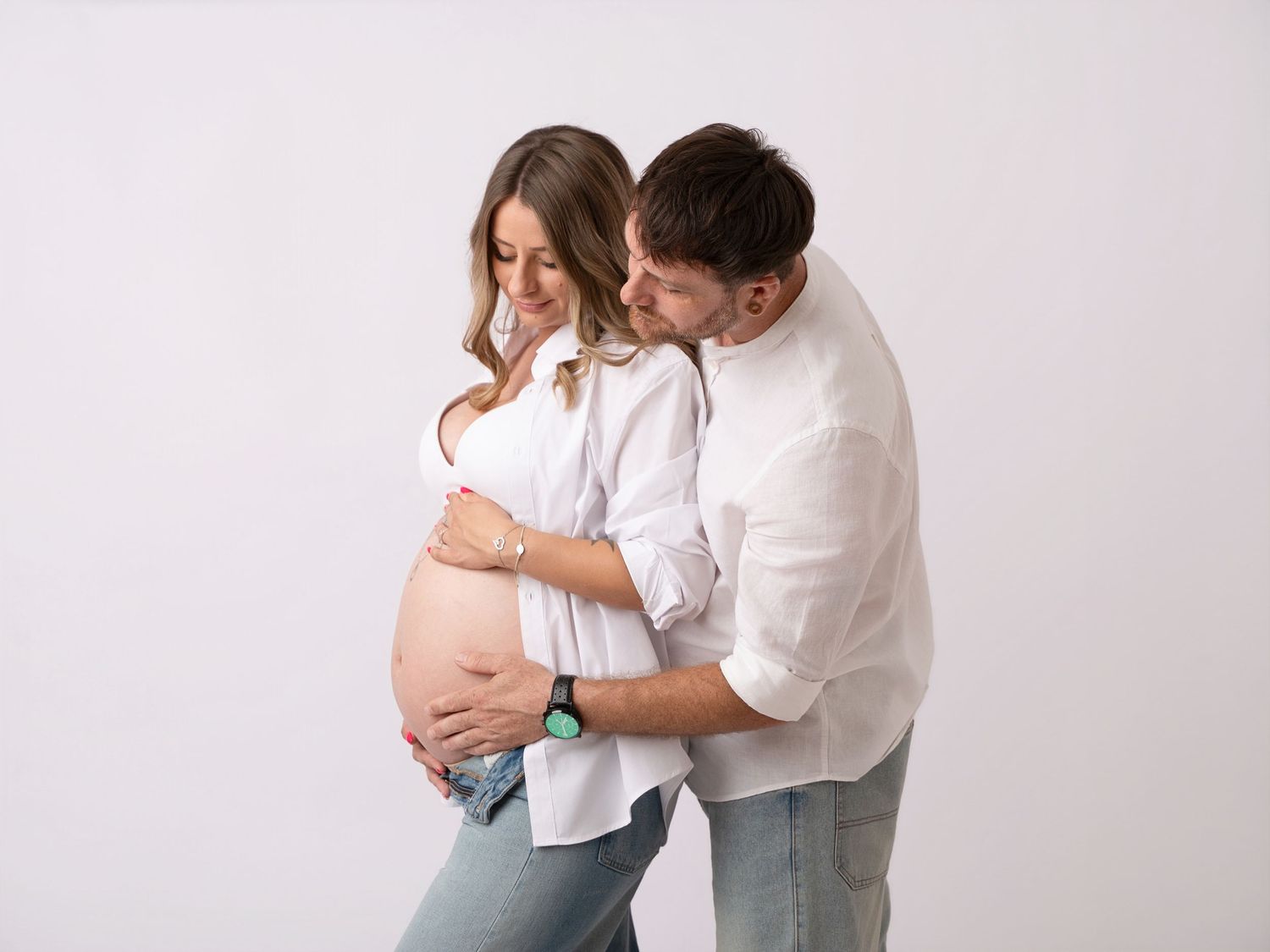 Couple sharing a tender maternity moment, man embracing pregnant woman from behind on white background.