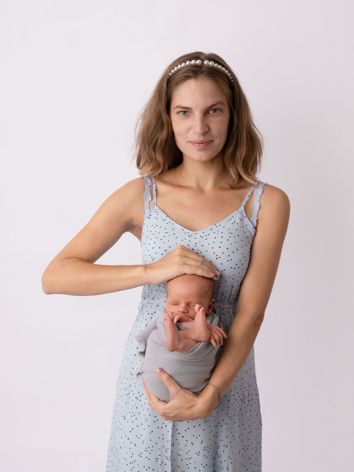 A woman in a light blue polka dot dress holds a swaddled newborn baby against a white background.
