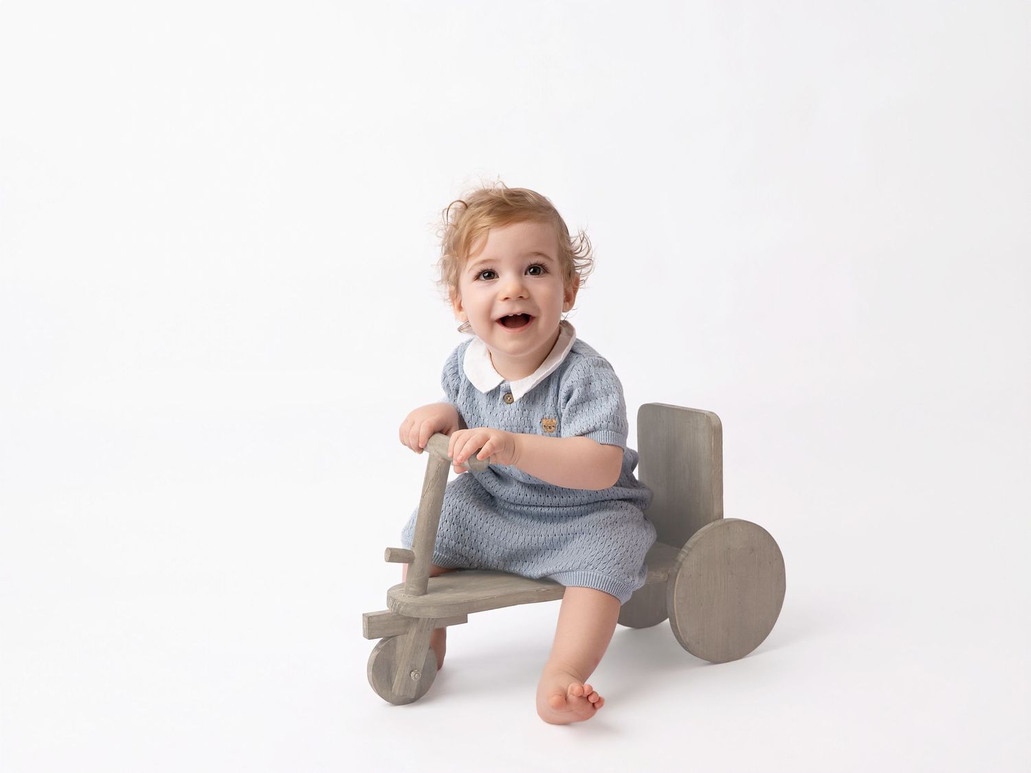 Smiling baby in blue knit romper sitting on a wooden toy tricycle against a white background.