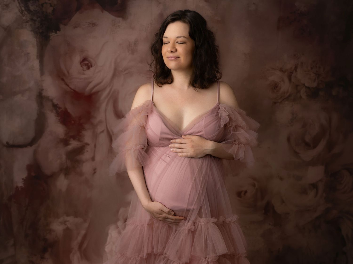 Pregnant woman in pink tulle maternity gown posing against a floral backdrop.