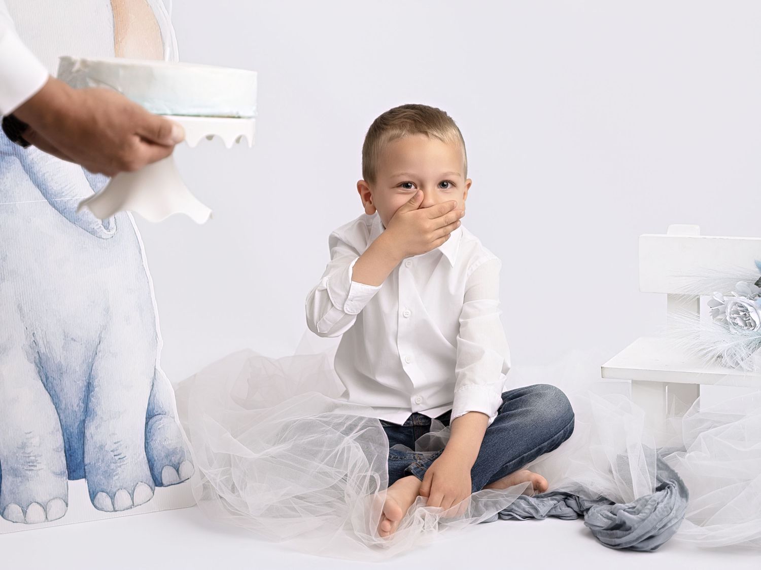 A young boy sits cross-legged on white floor, covering his mouth, beside tulle fabric and a white bench.