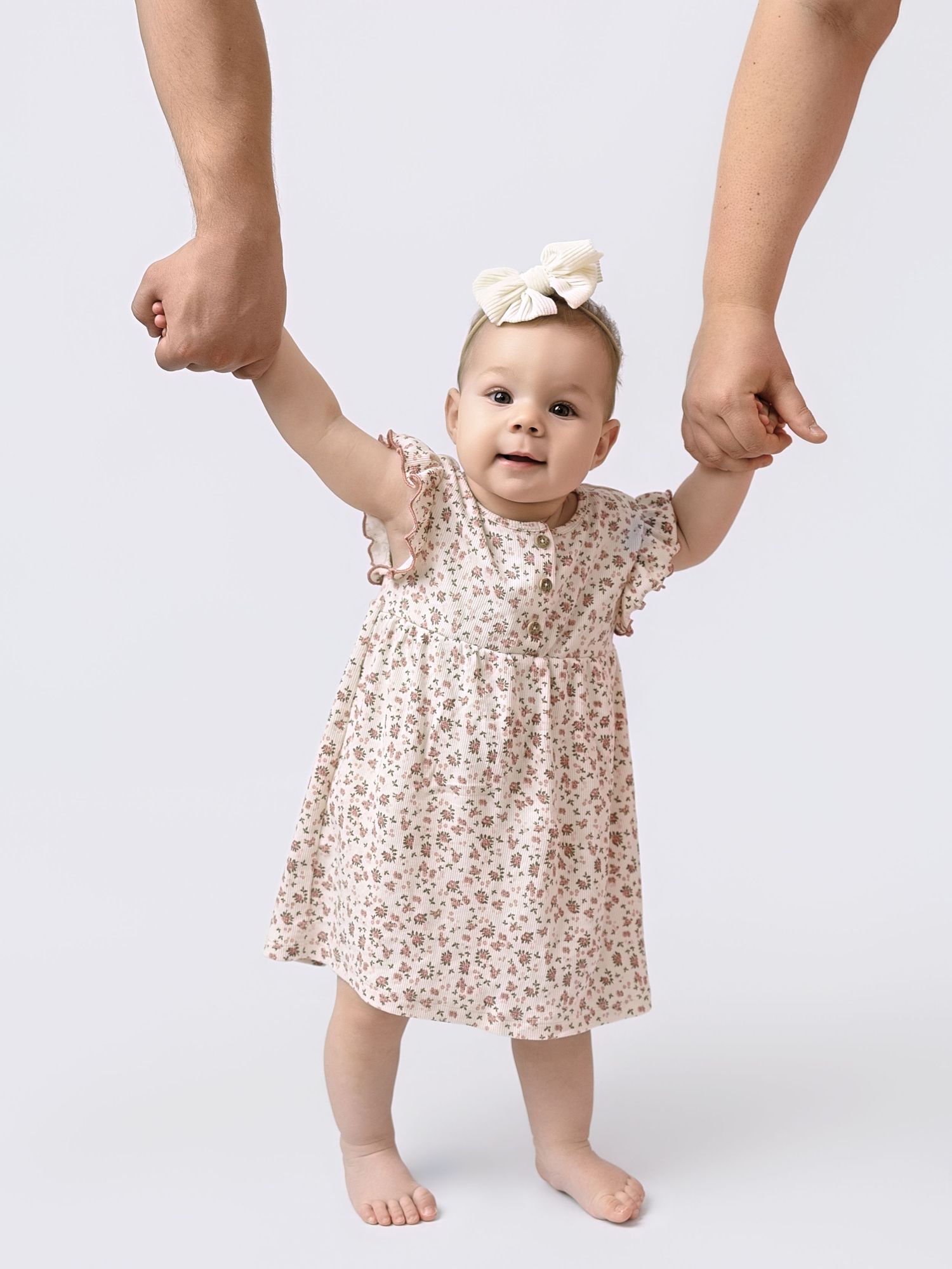 Happy baby girl in floral print dress and bow headband taking steps while being supported by two adults.