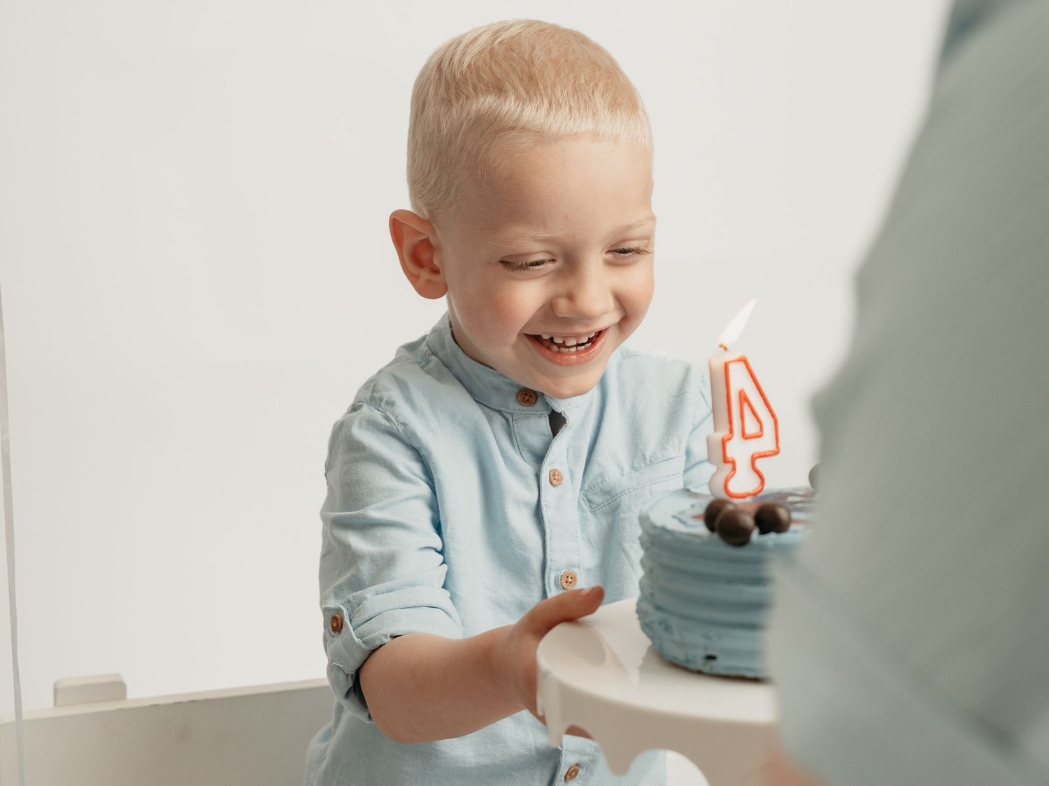 Smiling toddler boy celebrating 4th birthday with candle, wearing light blue shirt.