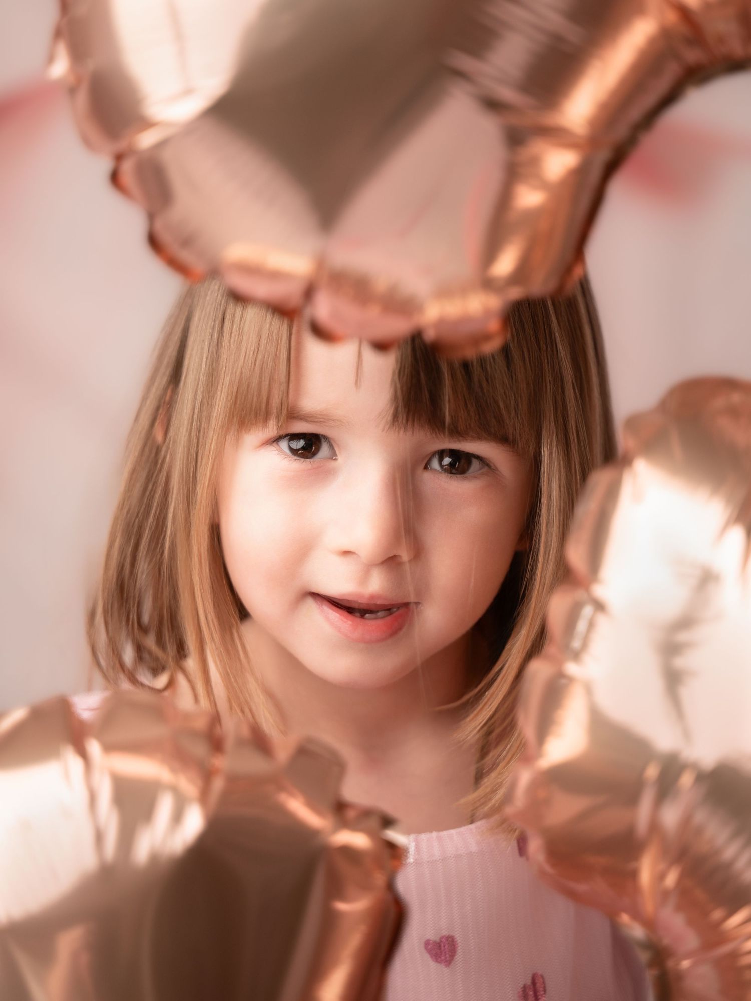 A young girl peeks through rose gold heart-shaped balloons in a close-up birthday portrait photo.