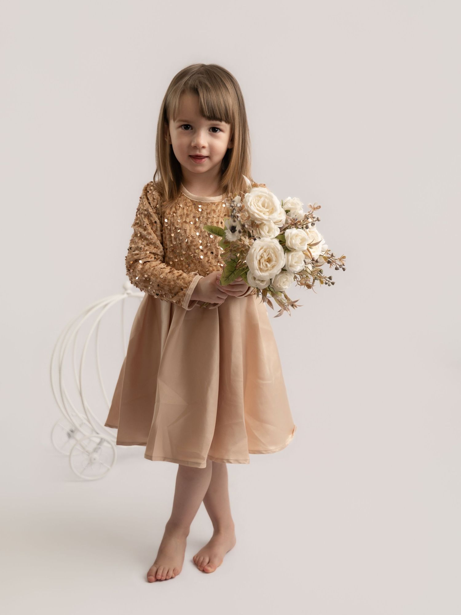A young girl in a gold sequin and champagne tulle dress holds a bouquet of white roses in a studio setting.