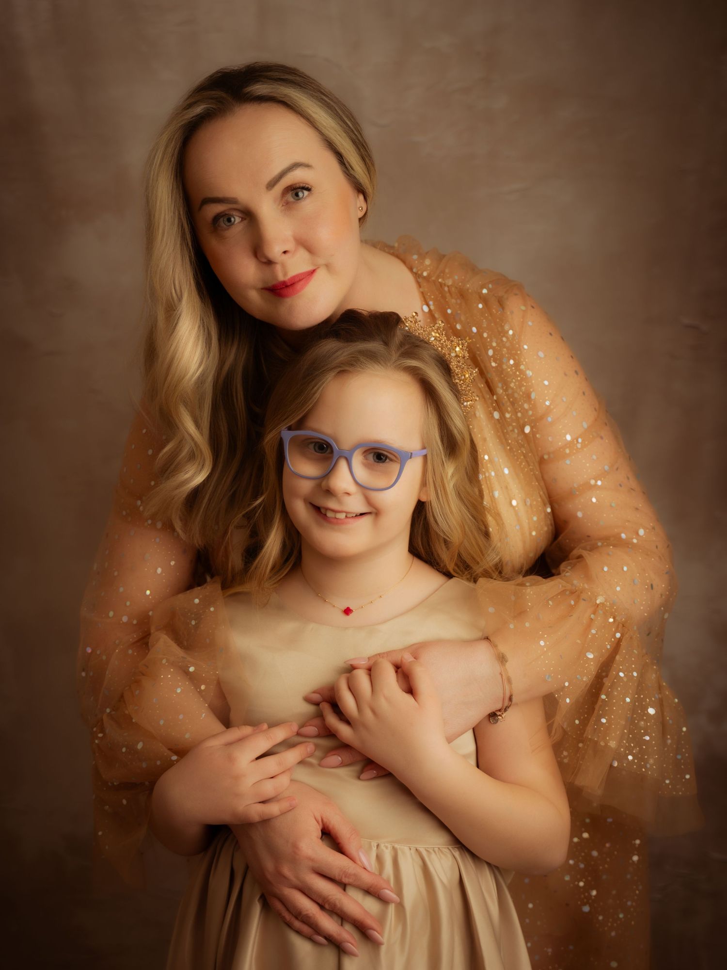 Mother in gold glitter dress embraces daughter in golden gown with blue glasses in studio.