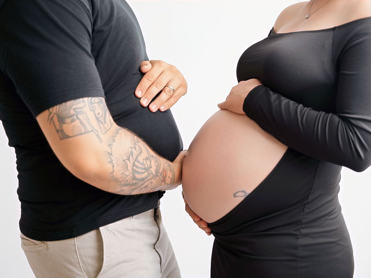 Pregnant couple in black outfits touching baby bumps together, man with tattoo sleeve, maternity photo shoot.