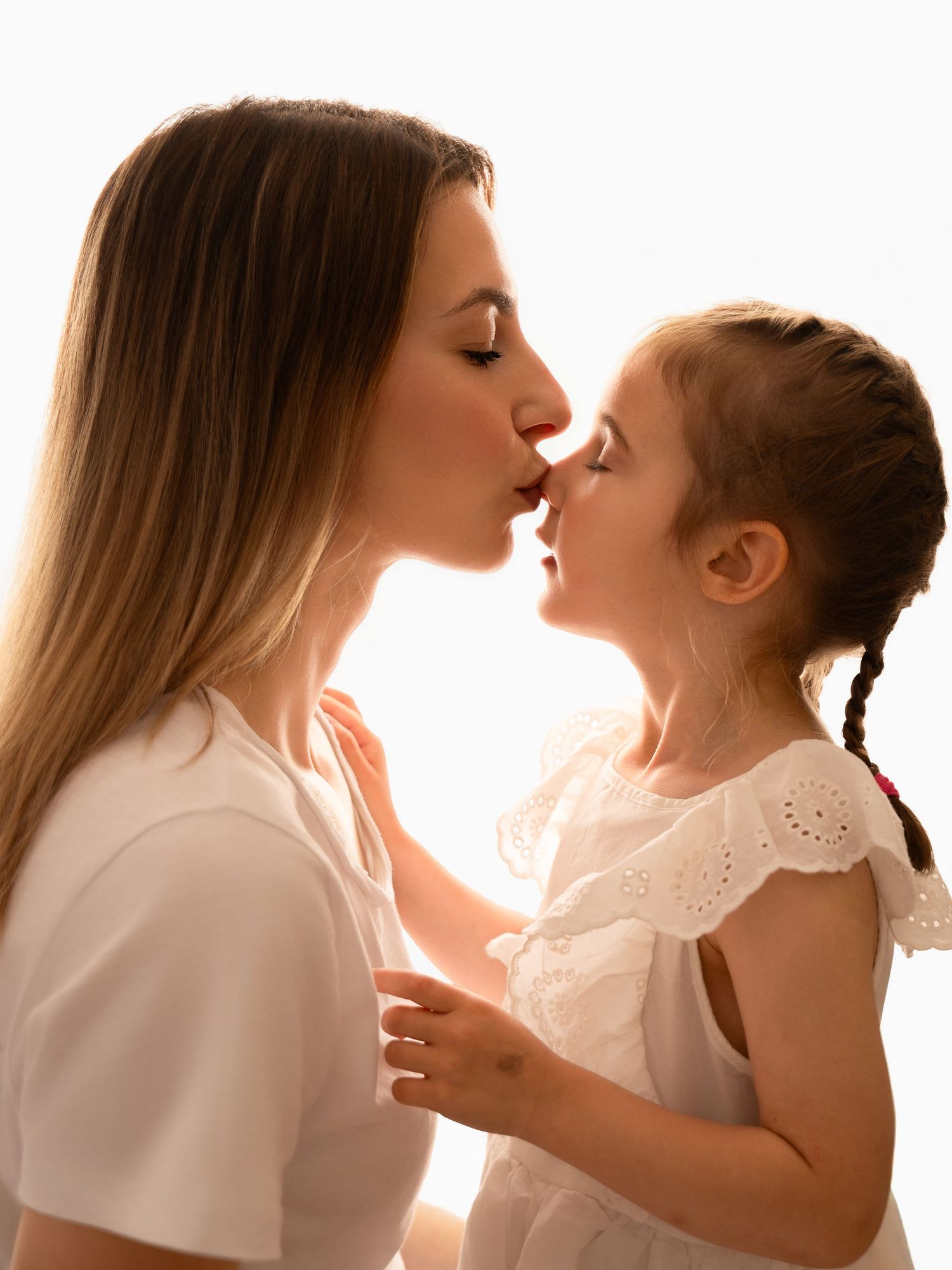 A mother and young daughter sharing a tender kiss, both wearing white, in a softly lit, heartwarming moment.