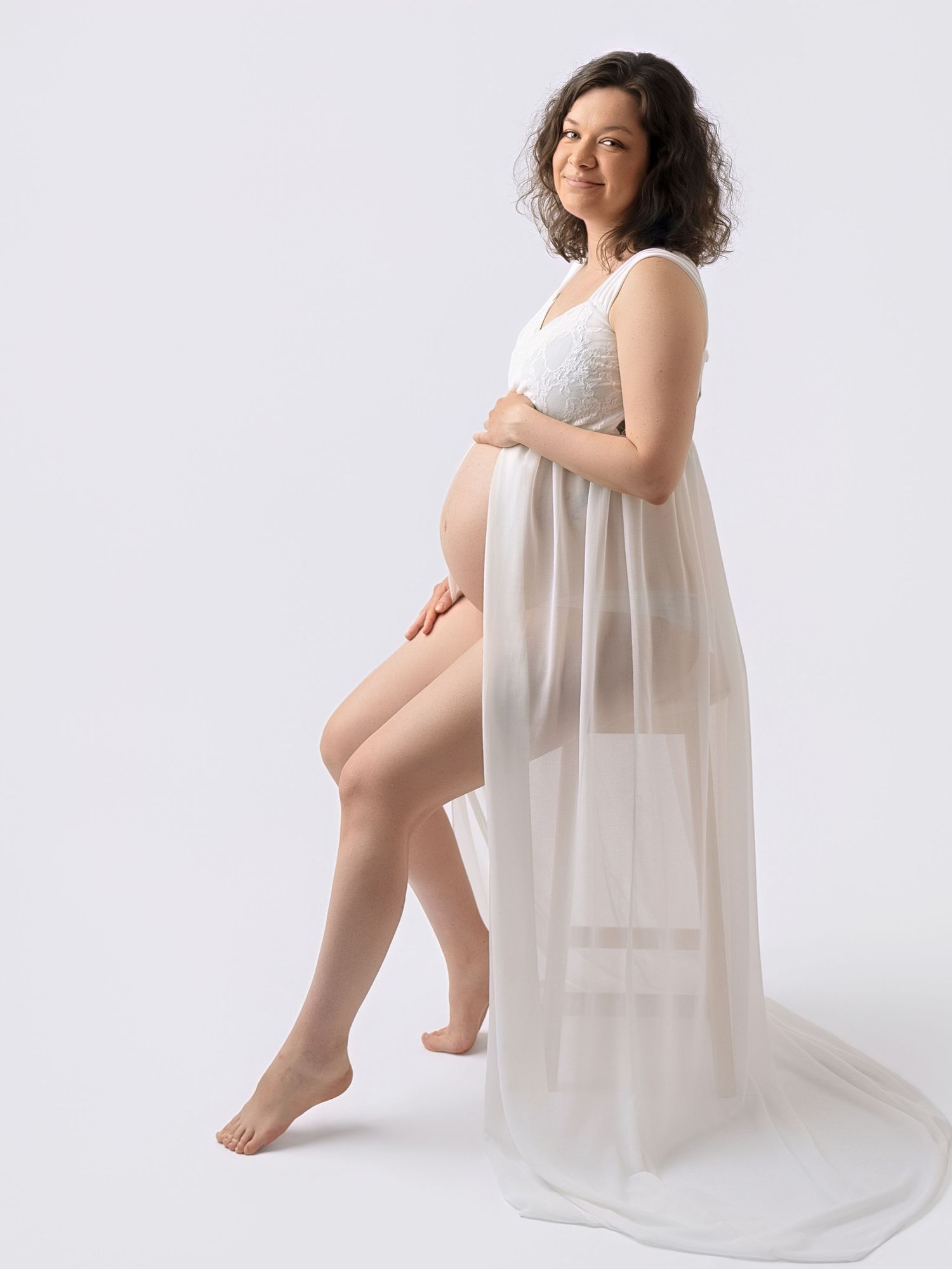 Pregnant woman in white lace bralette and sheer tulle skirt posing gracefully against a white background.