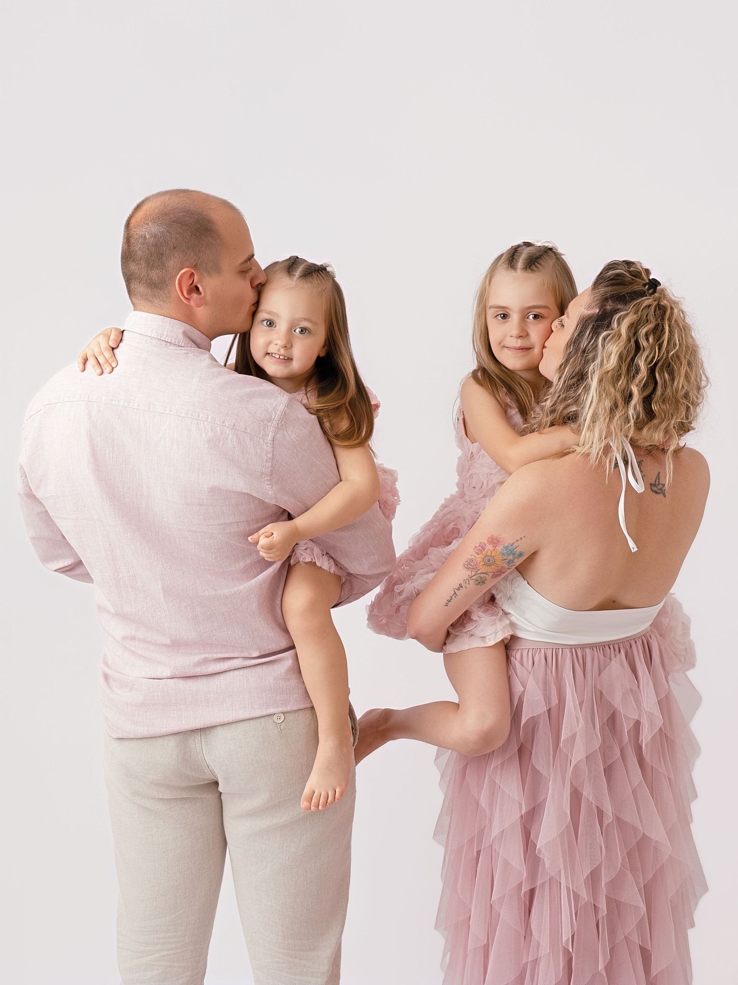 Family portrait of parents holding two young daughters in matching pink dresses against a white background.