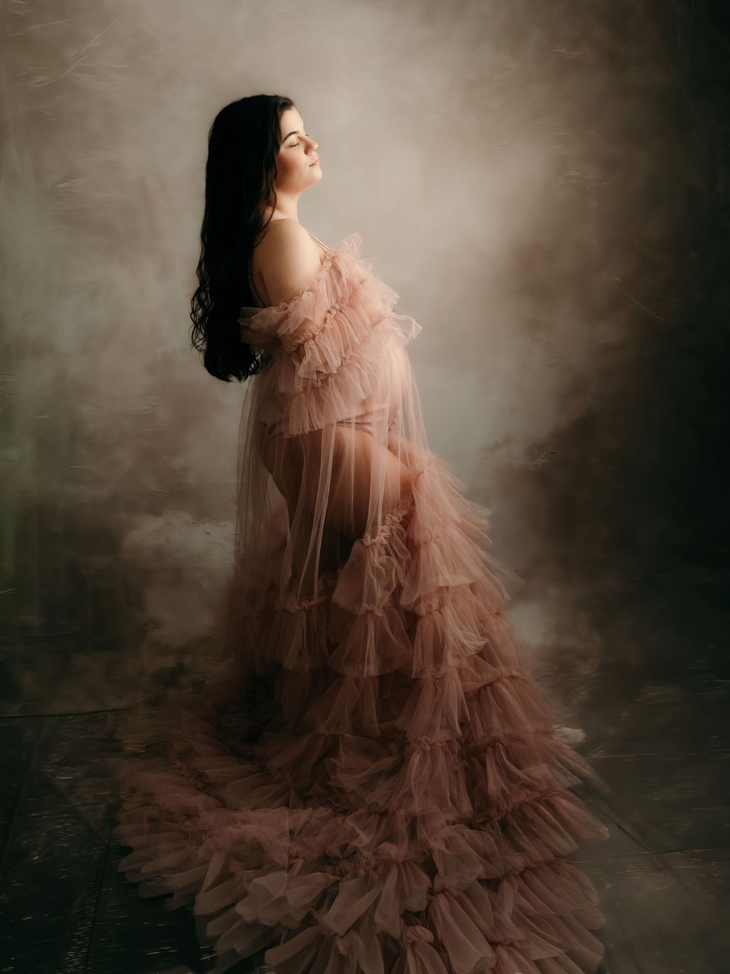 Pregnant woman in a dusty rose ruffled tulle maternity gown posing dramatically against a moody studio backdrop.