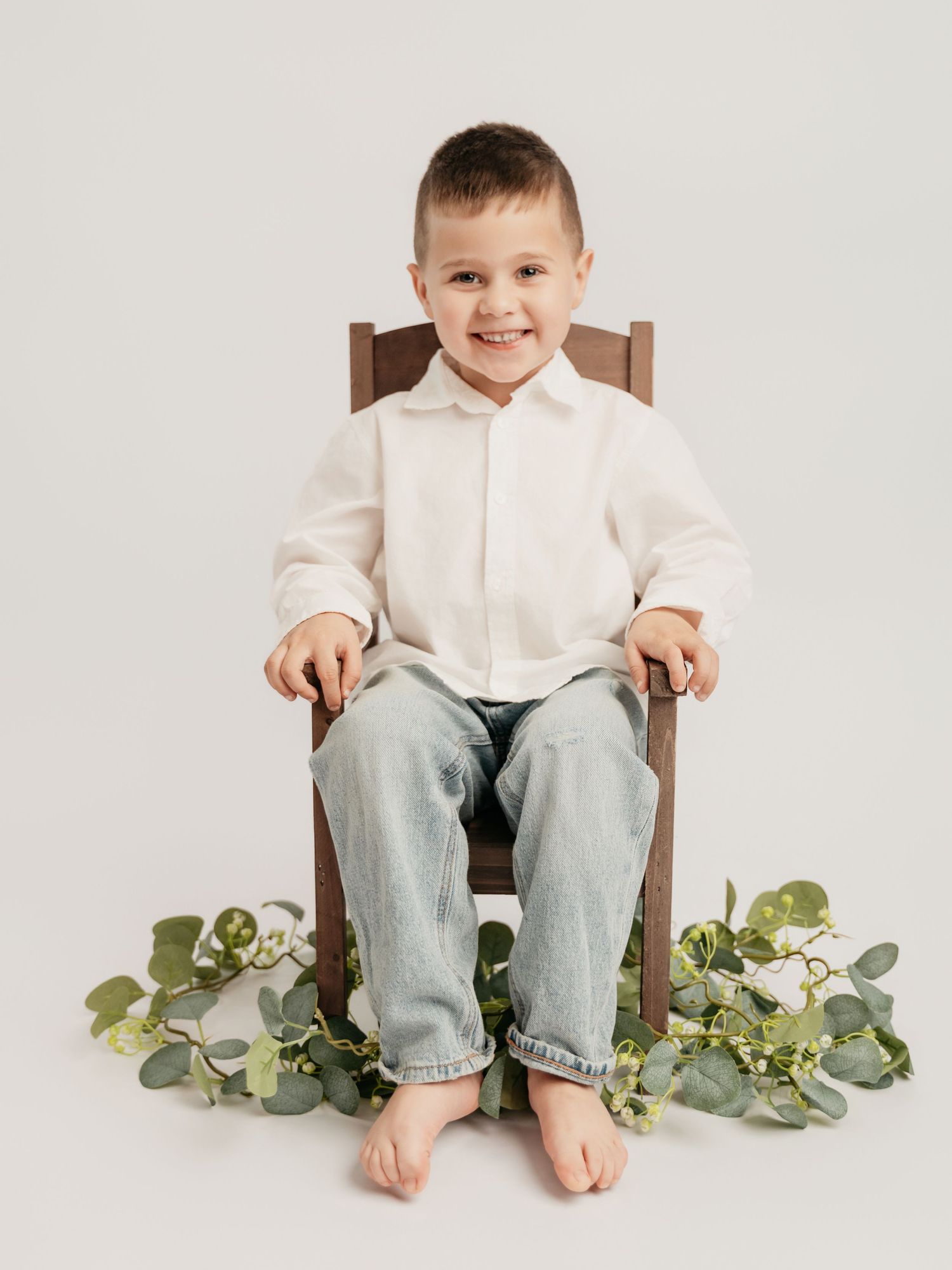 Smiling young boy in white shirt and jeans sitting on wooden chair with green eucalyptus decor.