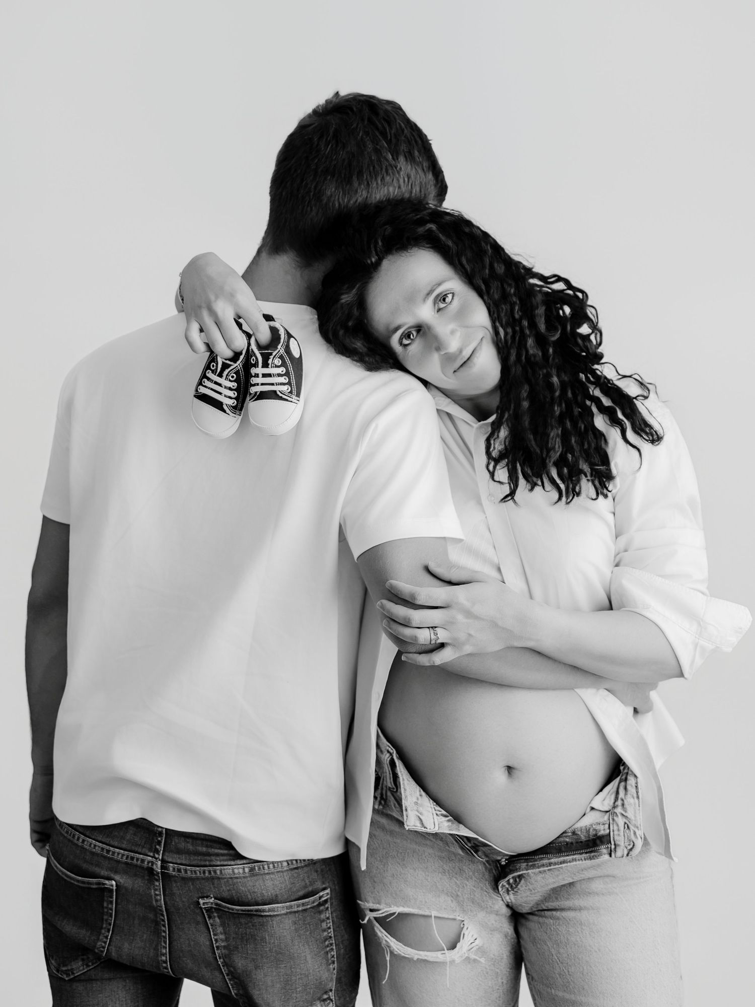 Black and white maternity photo of a couple holding tiny baby shoes while the pregnant woman rests her head on her partner.