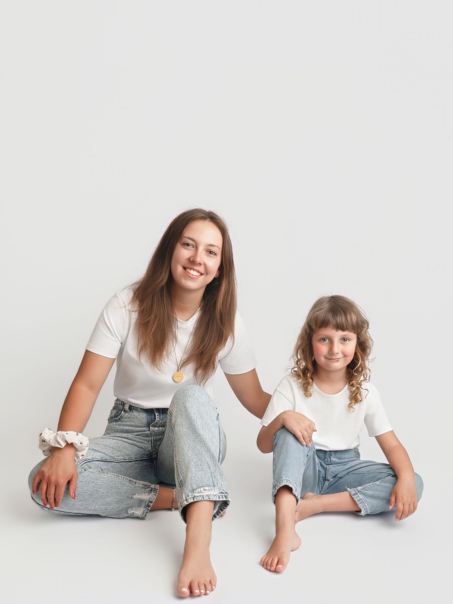Mother and daughter in matching white tees and jeans sit together smiling in bright studio photo.