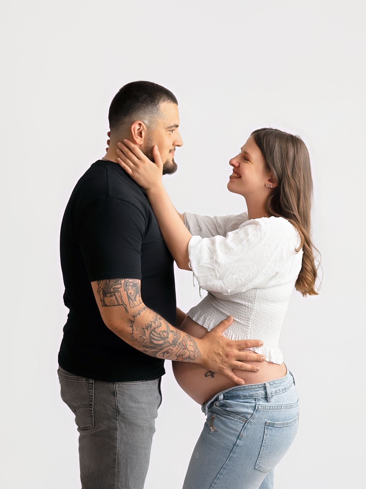 Couple sharing a tender moment, pregnant woman in white top and jeans with tattooed partner in black tee.