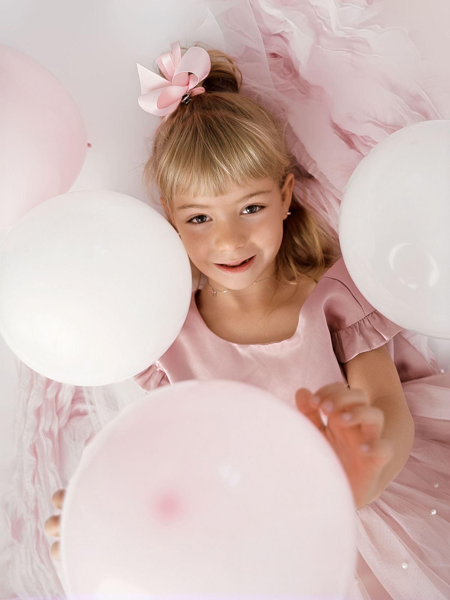 Young girl in pink dress holding white and pink balloons, wearing a pink bow hair accessory.