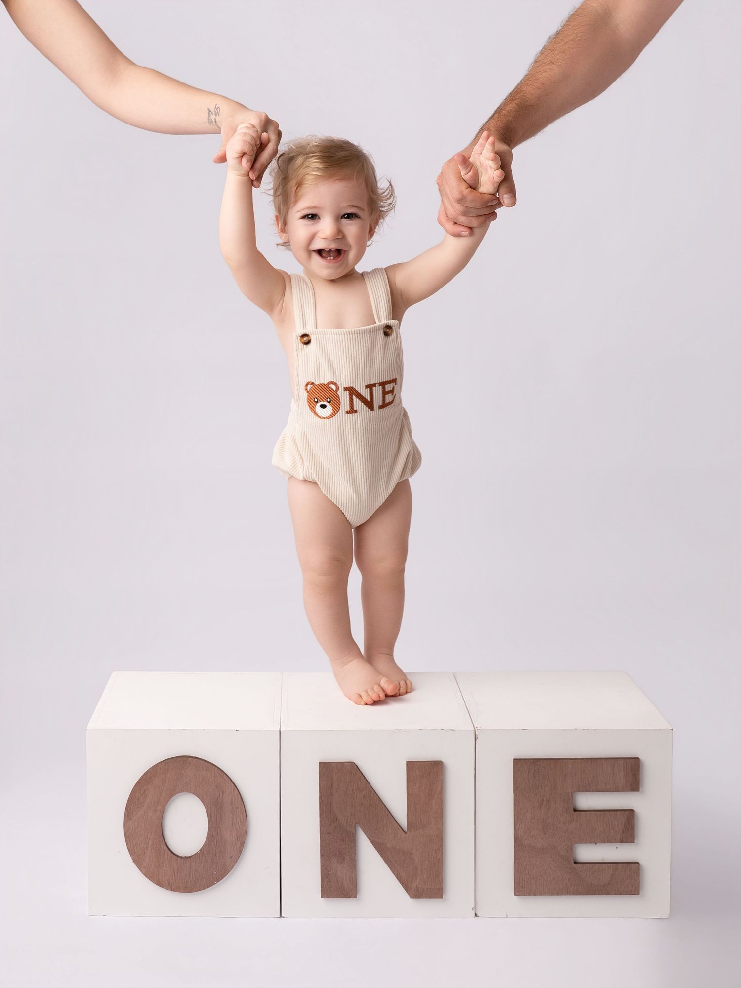 Smiling baby standing on ONE letter blocks held by parents hands in birthday photoshoot.