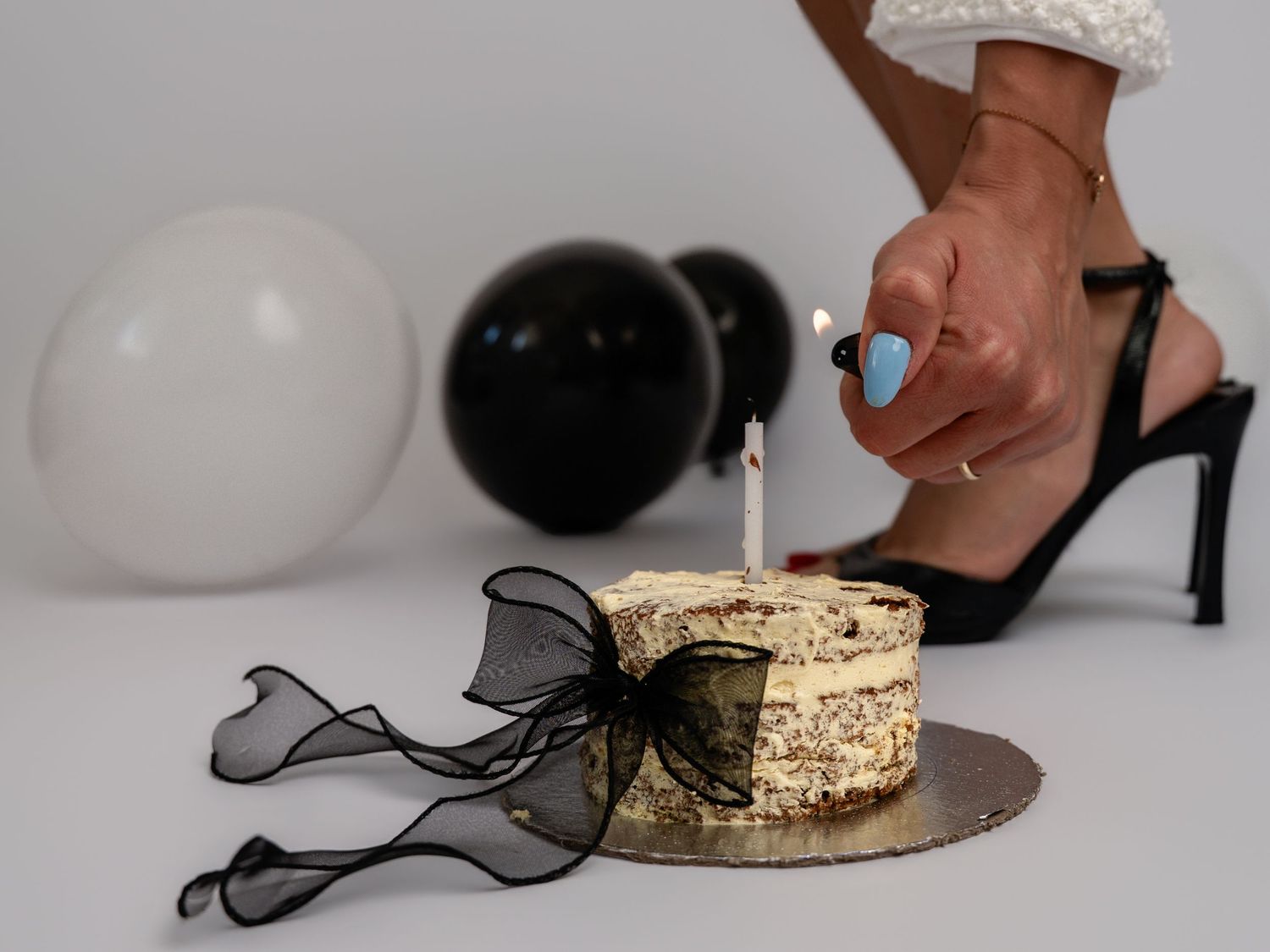 A person lights a candle on a decorated birthday cake surrounded by black and white balloons in a studio setting.