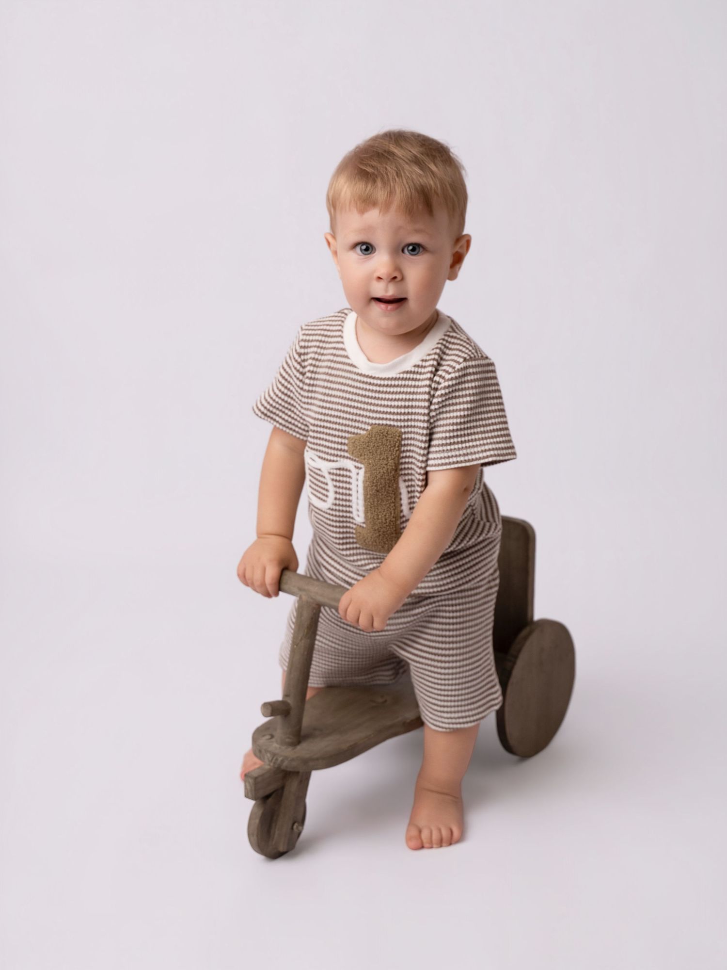 Toddler boy in striped brown romper sitting on wooden balance bike toy against white background.