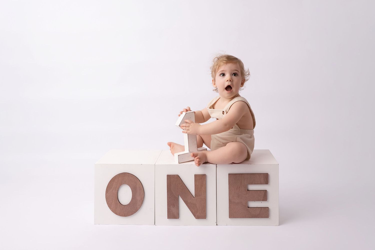 Baby in beige onesie sits on white ONE blocks celebrating first birthday in a clean studio photo.