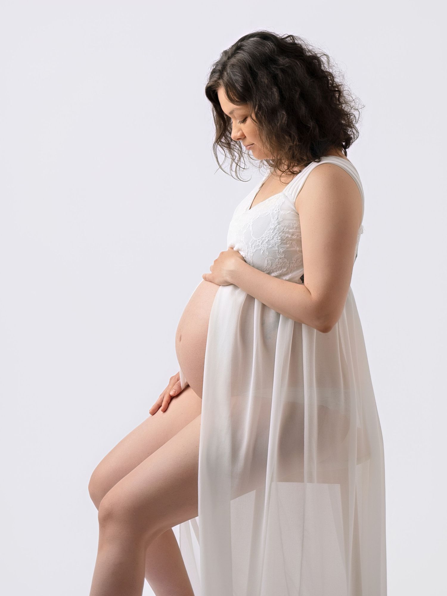 Pregnant woman in white lace and sheer gown sitting gracefully, cradling baby bump in professional studio photo.
